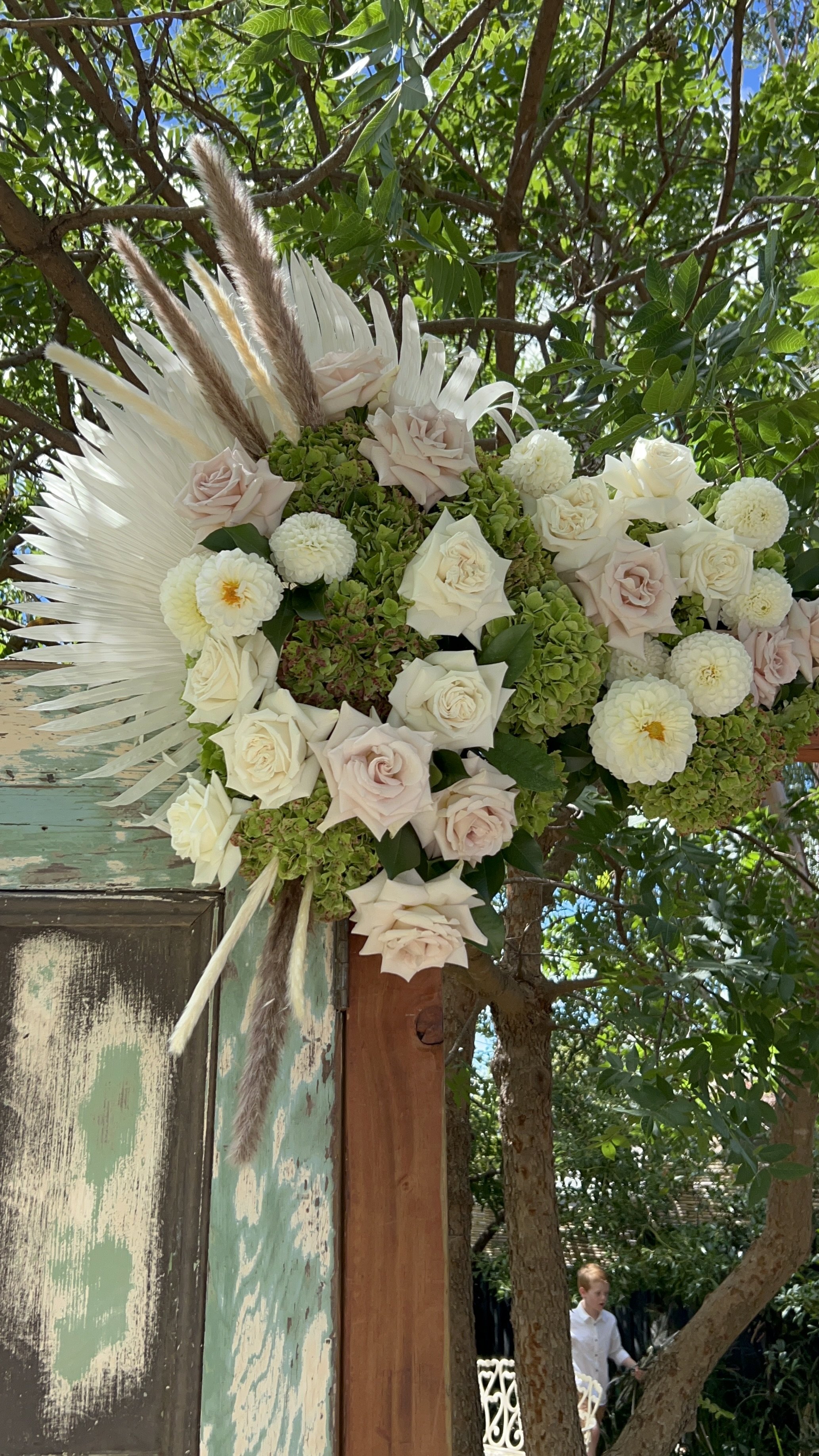 A large floral arrangement with white and pale pink roses, white dahlias, and green hydrangeas, accented with pampas grass, attached to a rustic wooden structure outdoors with trees in the background.