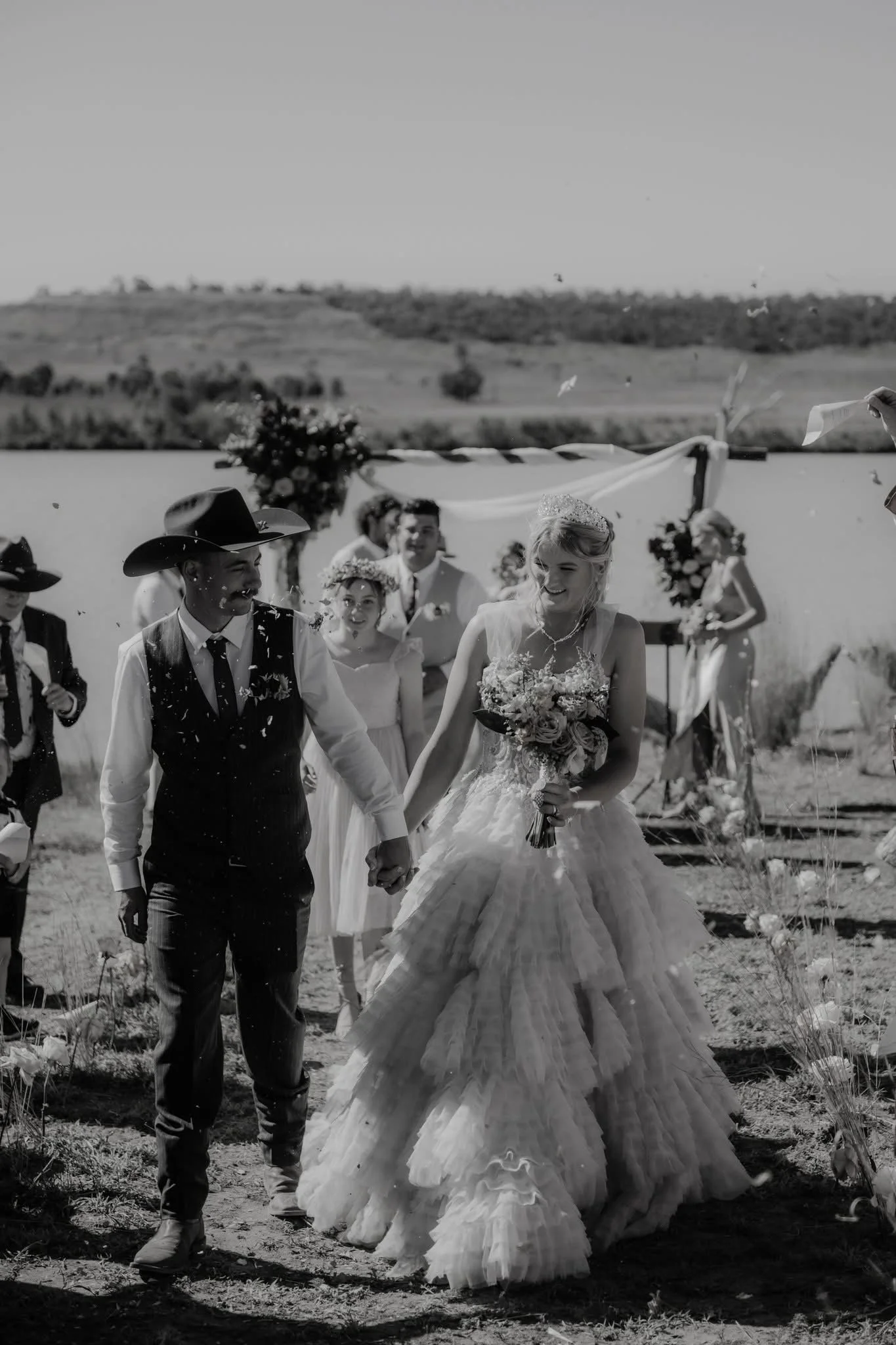 Black and white photo of a wedding ceremony outdoors, featuring a smiling bride in a wedding gown and a groom in a vest and cowboy hat holding hands, surrounded by guests and a scenic rural background.