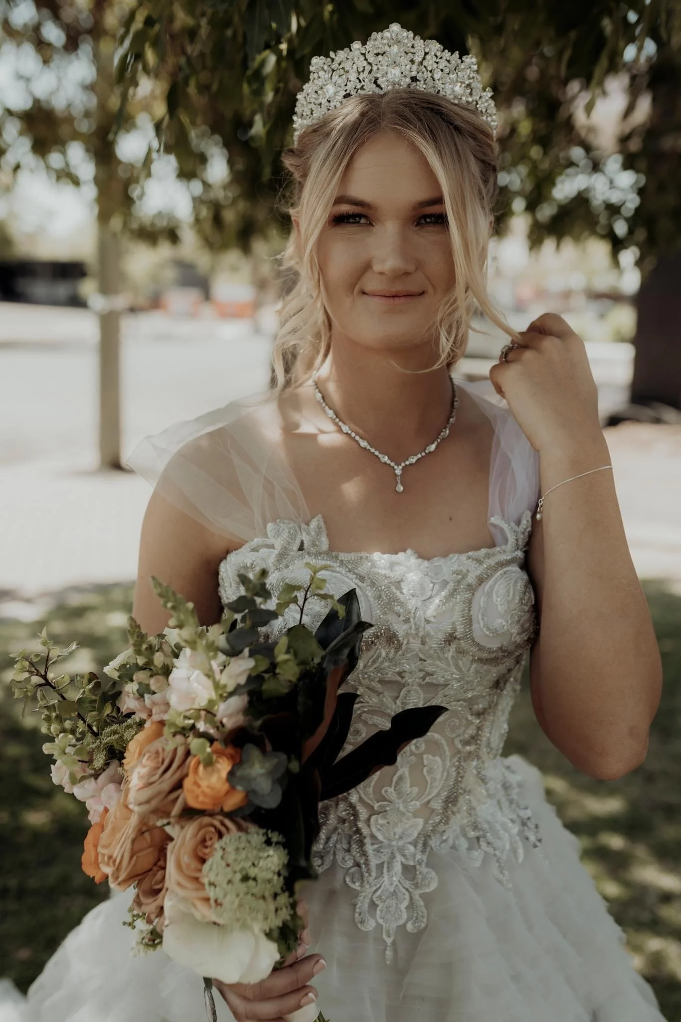 A bride with blonde hair, wearing a white lace wedding gown and a pearl necklace, holding a bridal bouquet with peach, white, and green flowers. She wears a tiara and poses outdoors under a tree, smiling softly.