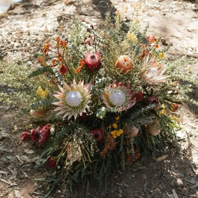 A floral arrangement with proteas, apples, and various wildflowers on the ground outdoors.