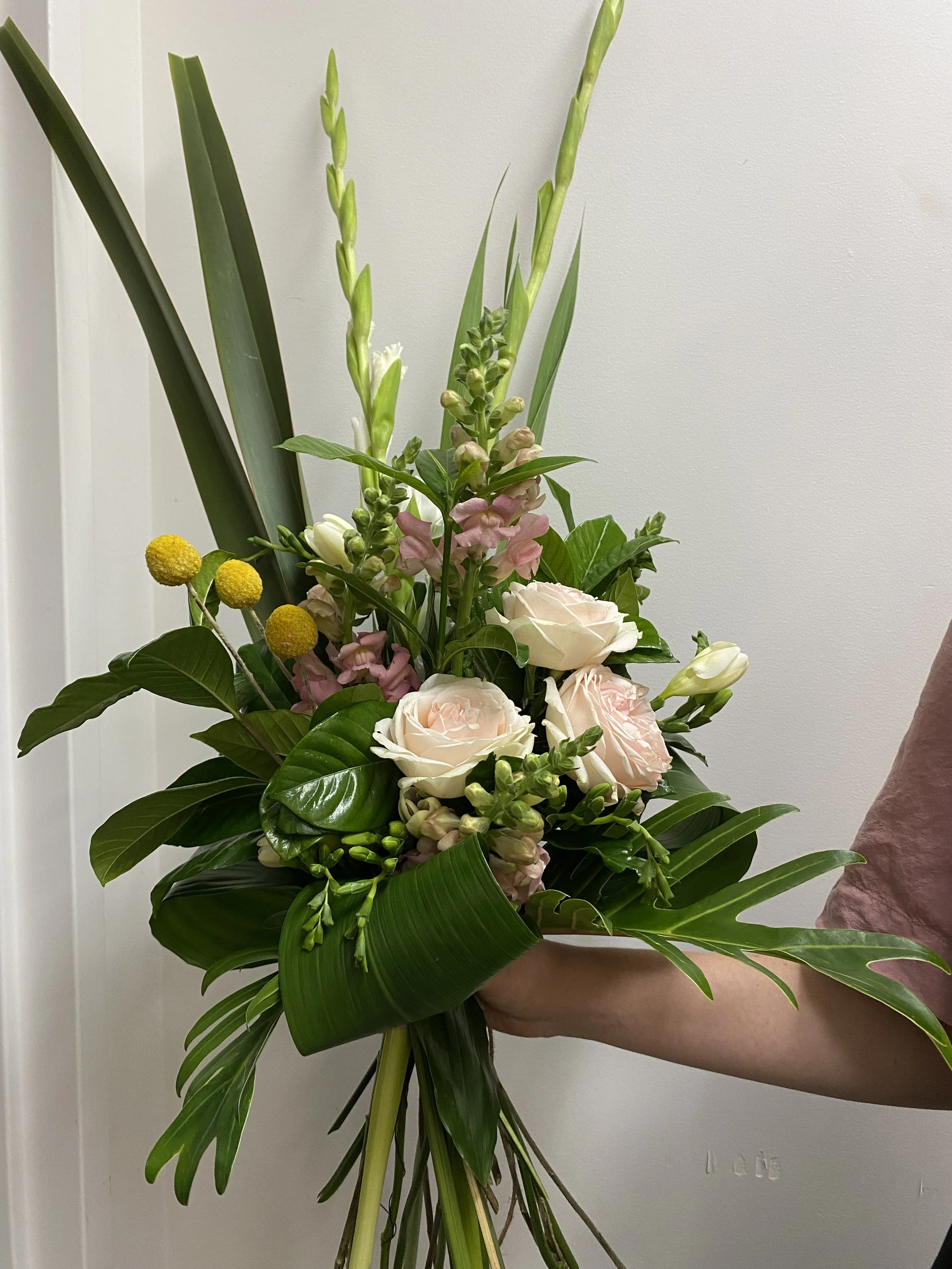 A person holding a colorful bouquet of flowers, including white roses, pink flowers, and yellow spherical flowers, with large green leaves, against a neutral background.