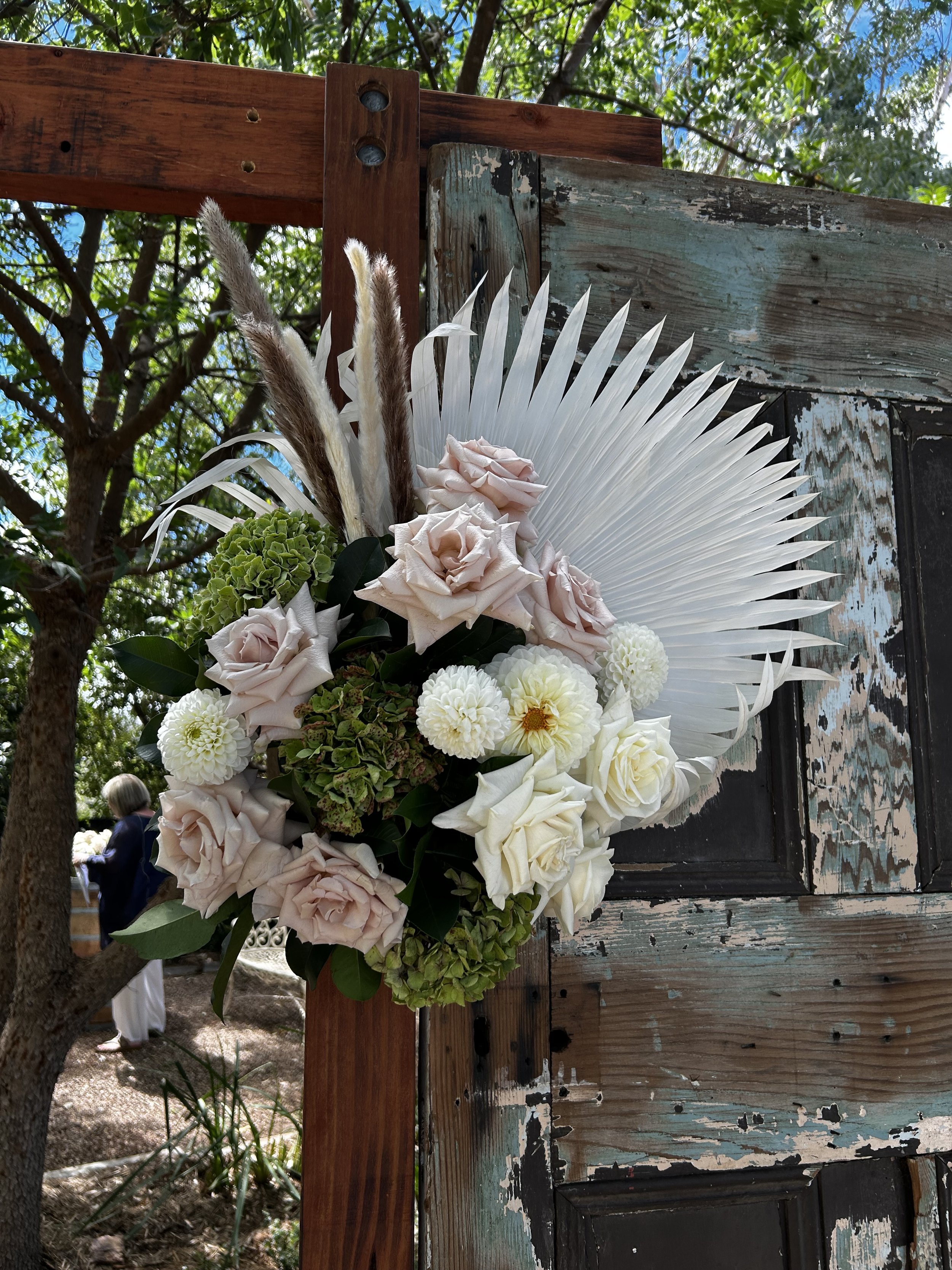 A floral arrangement featuring white and pale pink roses, white dahlias, green hydrangeas, and decorative pampas grass, with a large white palm leaf backdrop mounted on a rustic wooden surface outdoors.