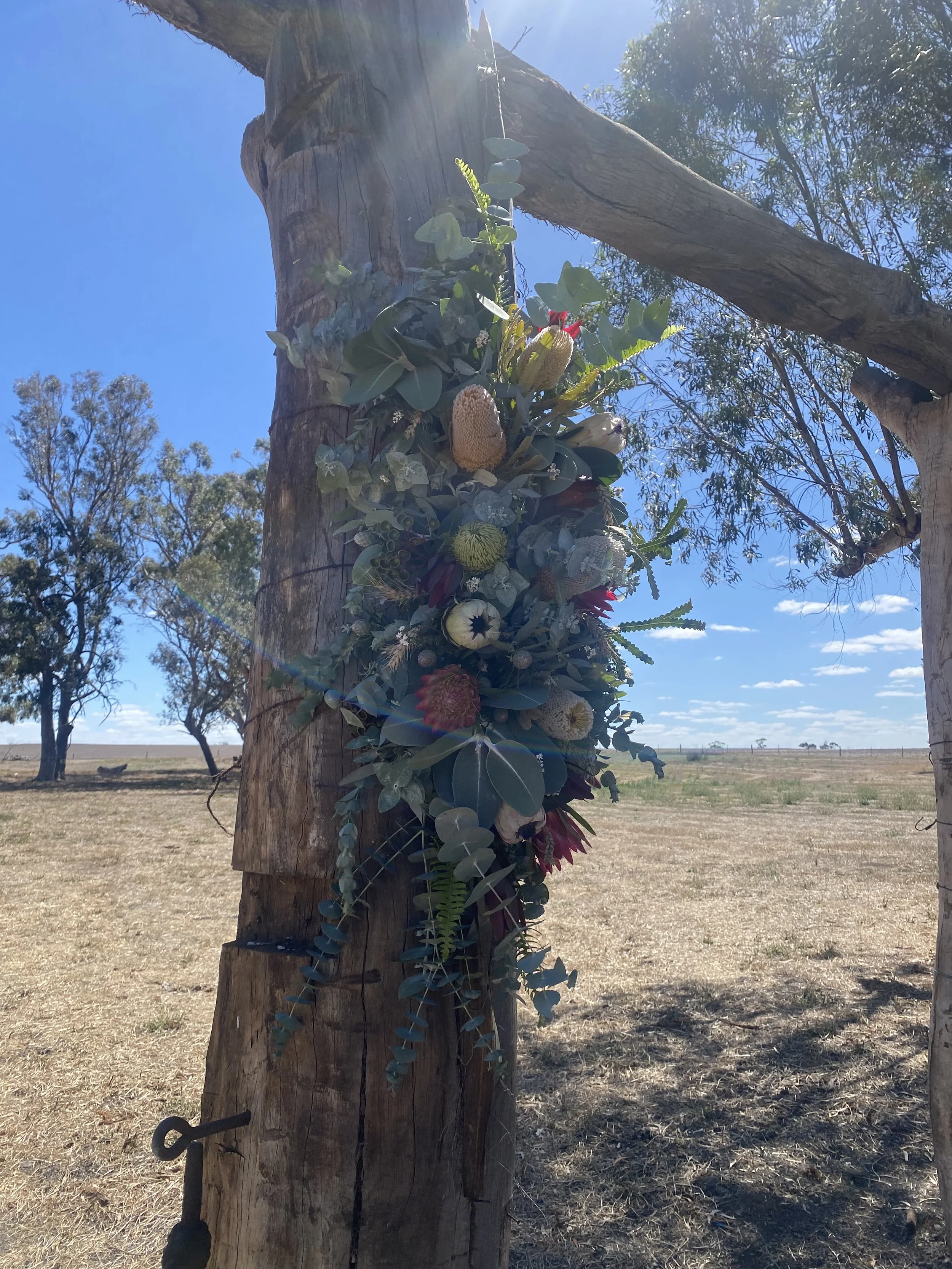 A flower arrangement attached to a wooden post outdoors on a bright, sunny day with a blue sky and sparse trees in the background.