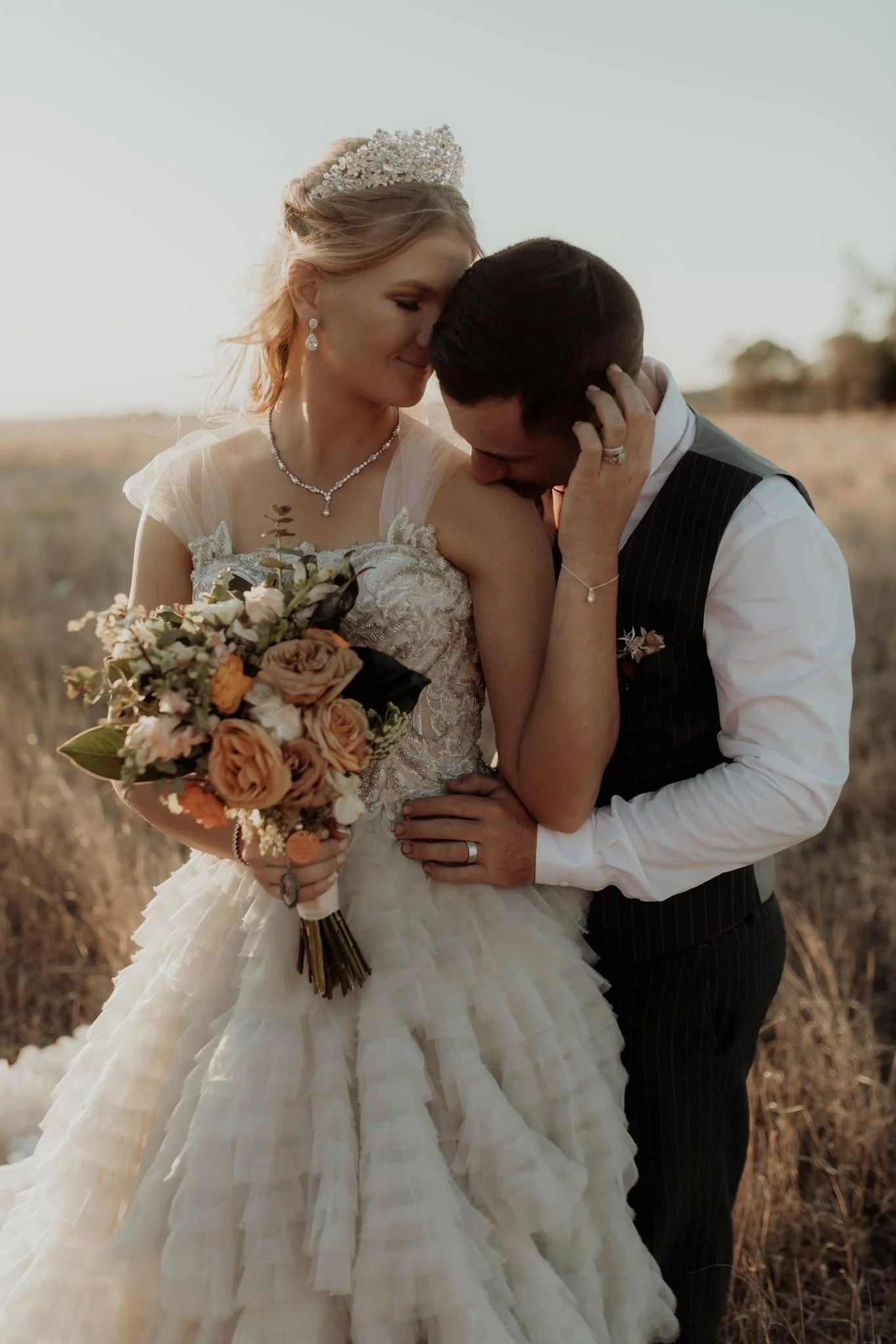 A bride and groom sharing a tender moment outdoors during sunset, with the bride holding a bouquet of peach, white, and orange flowers. The bride is wearing a detailed lace wedding gown and a tiara, and the groom is dressed in a black pinstripe vest and white shirt.