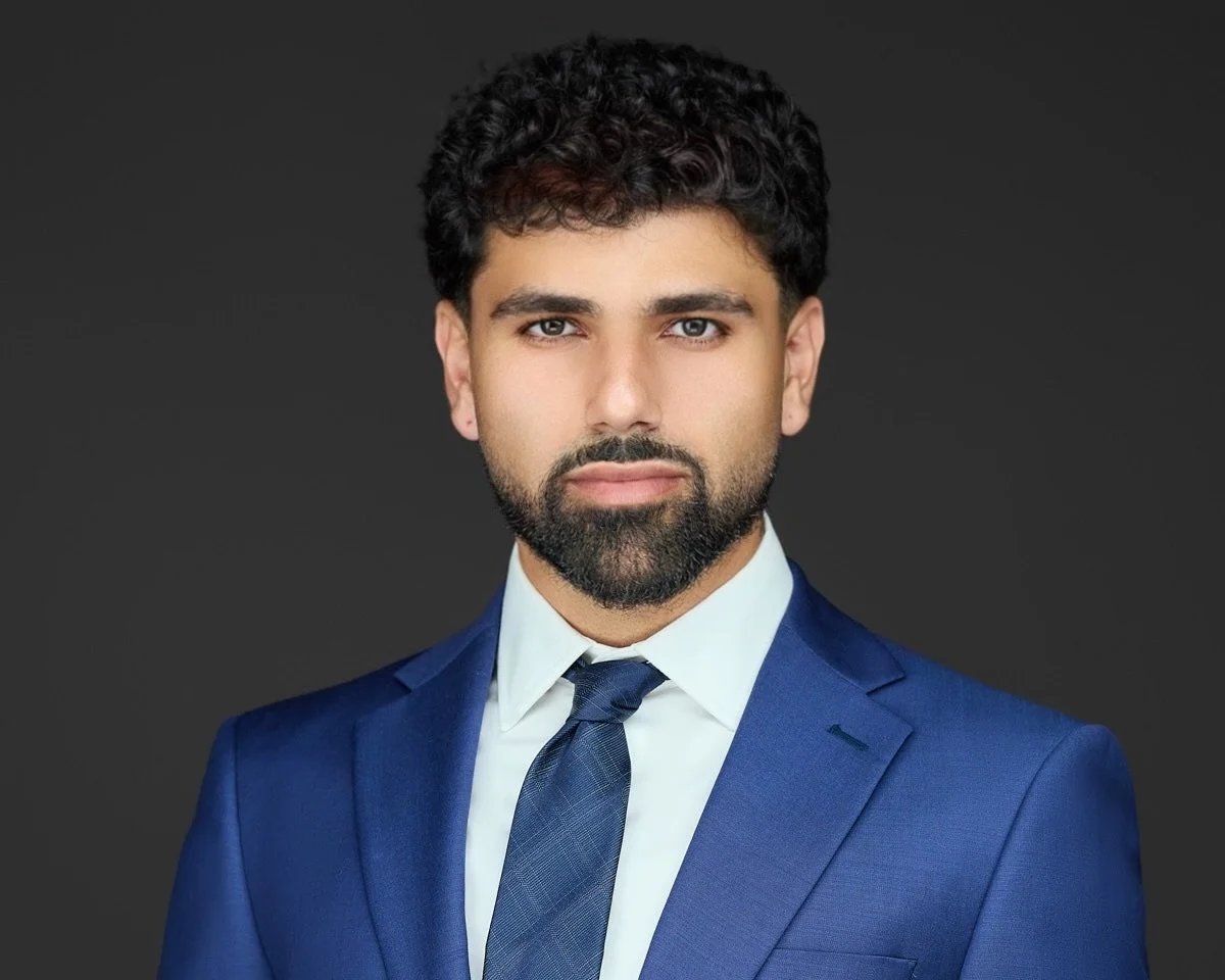 Headshot of a man with dark curly hair and a beard, dressed in a navy blue suit, white shirt, and blue tie, against a dark gray background.