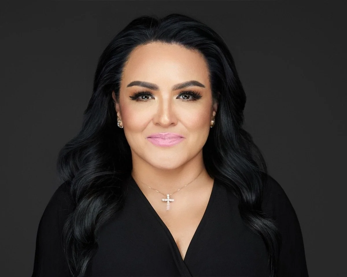 Portrait of a woman with long black hair, light makeup, wearing a black top, pearl earrings, and a cross necklace, against a dark background.