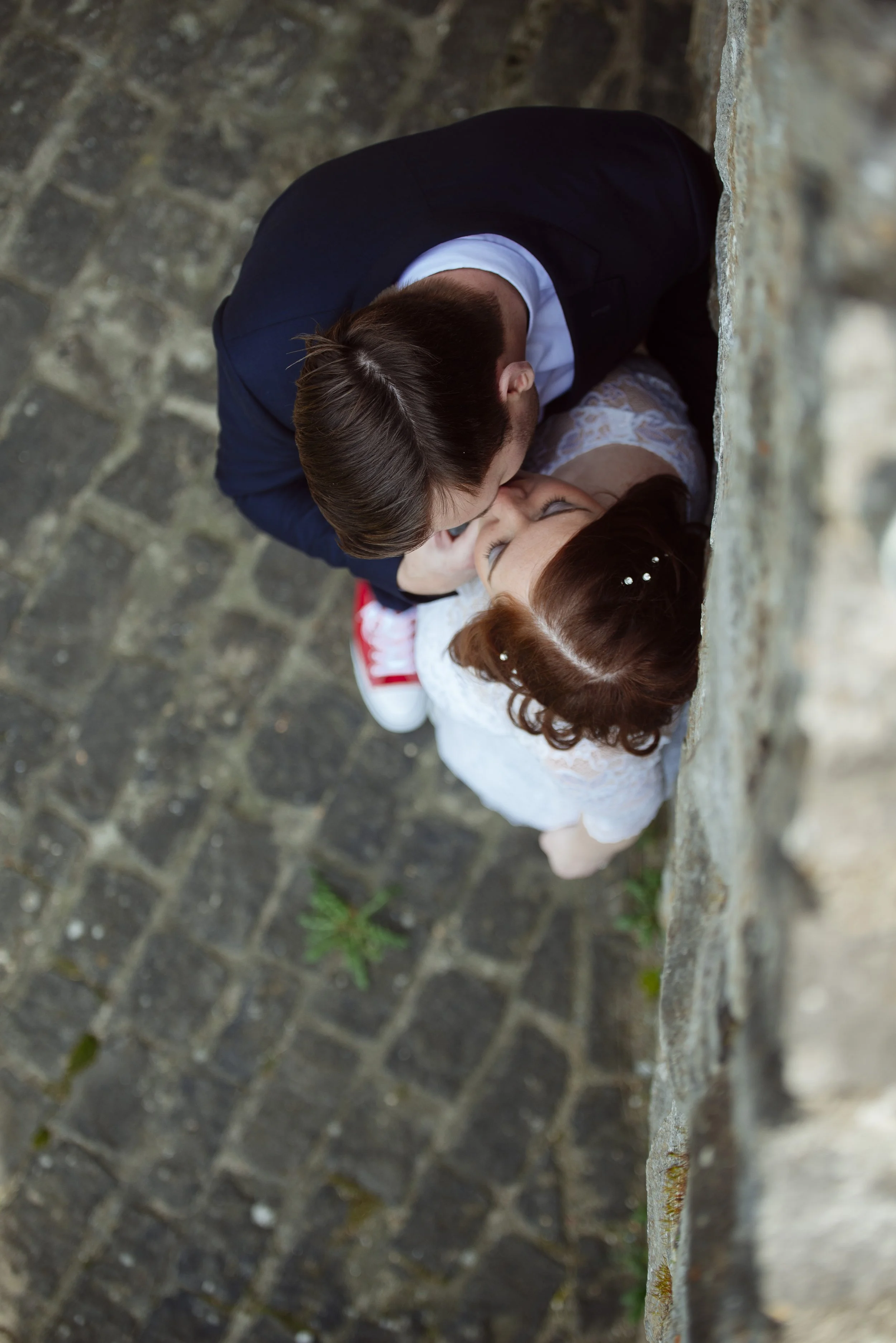 Ein Mann und eine Frau küssen sich in einer Gasse, die mit Kopfsteinpflaster bedeckt ist, hinter einer Steinmauer.