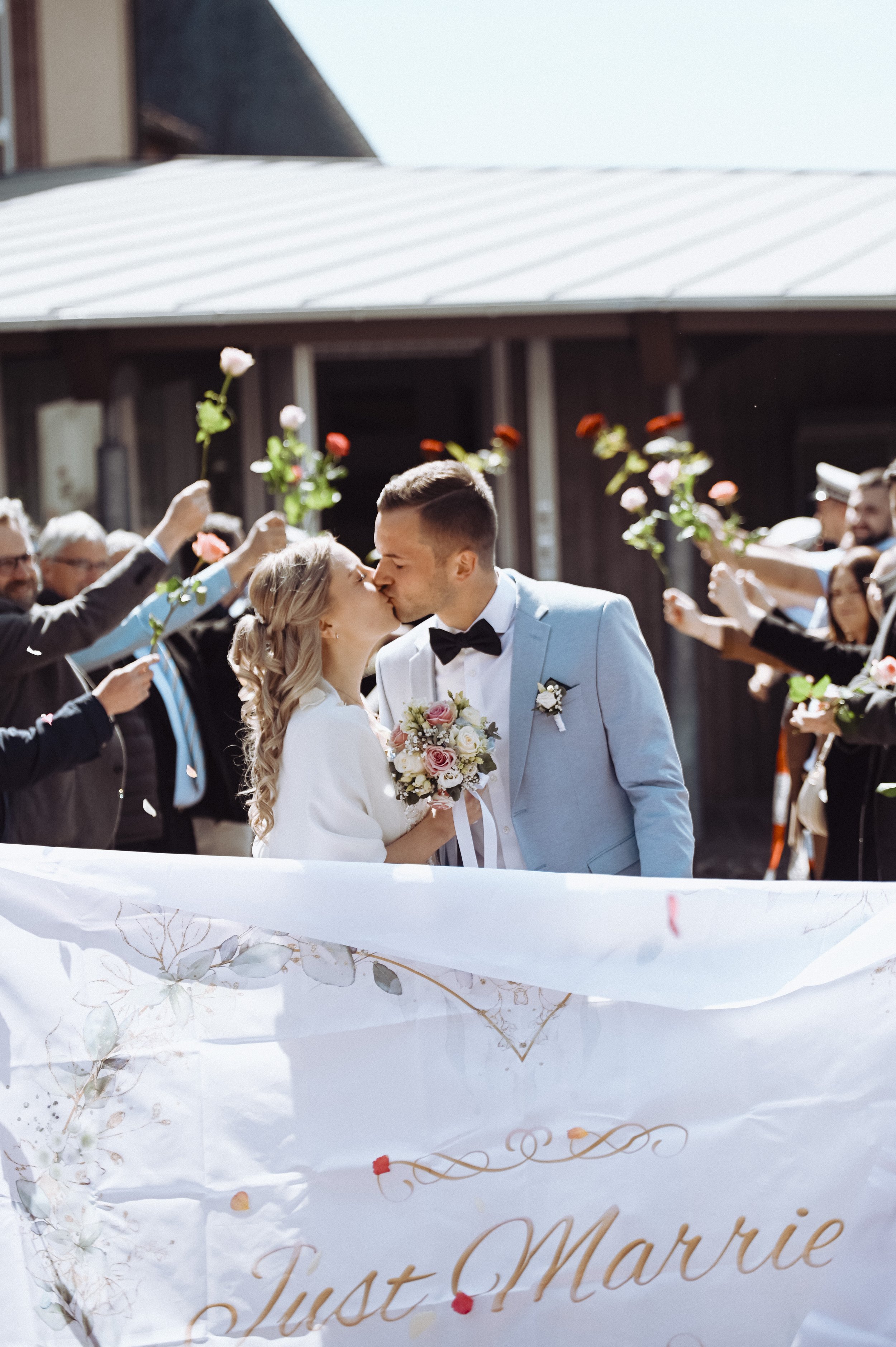 Ein frisch verheiratetes Paar, das sich küsst, umgeben von Freunden und Familie, die Rosen und Blumen für die Glückwünsche halten, bei einer Hochzeitsfeier im Freien.