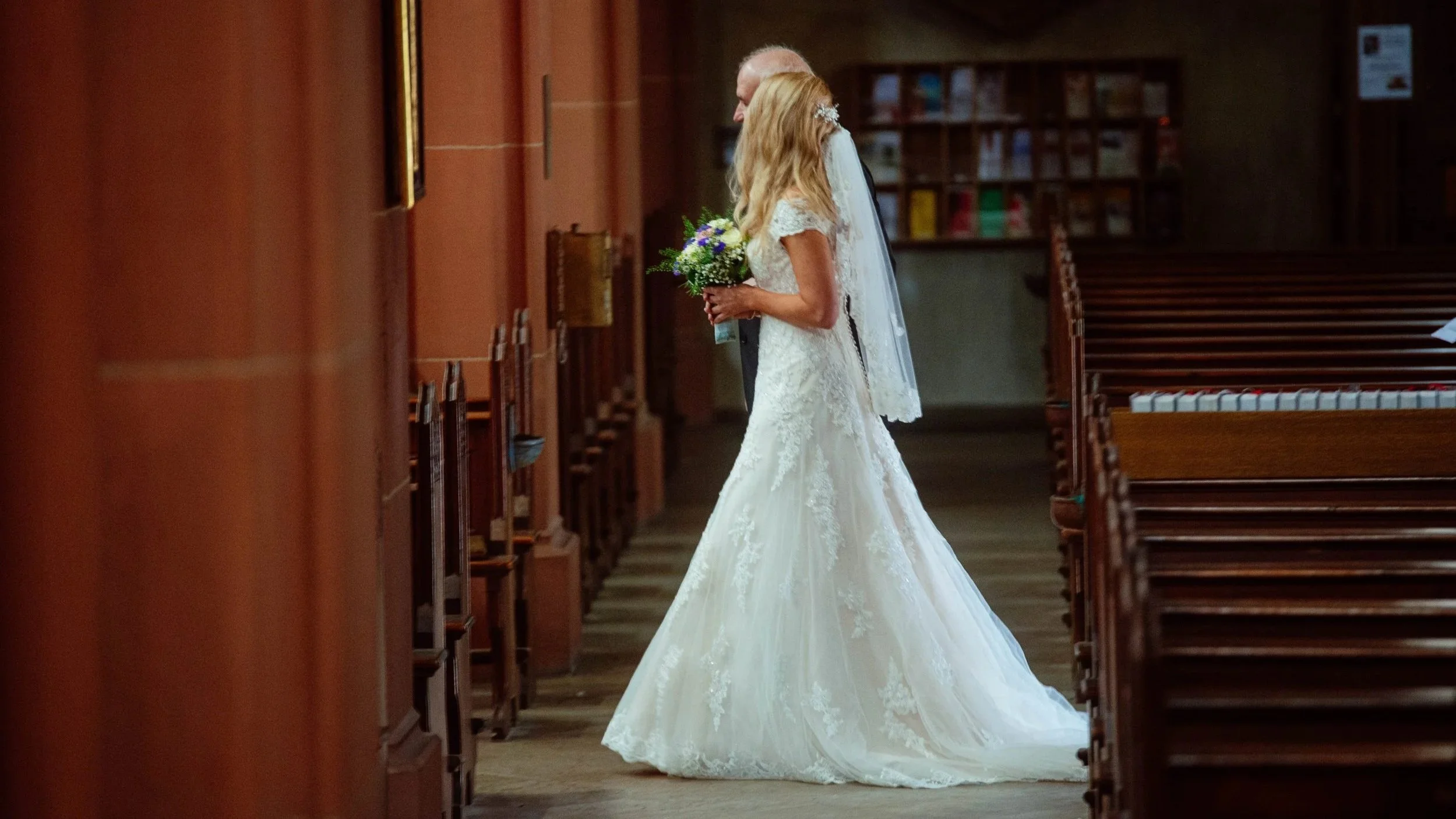 Eine Hochzeitsszene in einer Kirche, ein Brautpaar steht sich gegenüber, die Braut trägt ein weißes Hochzeitskleid und hält einen Blumenstrauß, während der Bräutigam einen dunklen Anzug trägt.