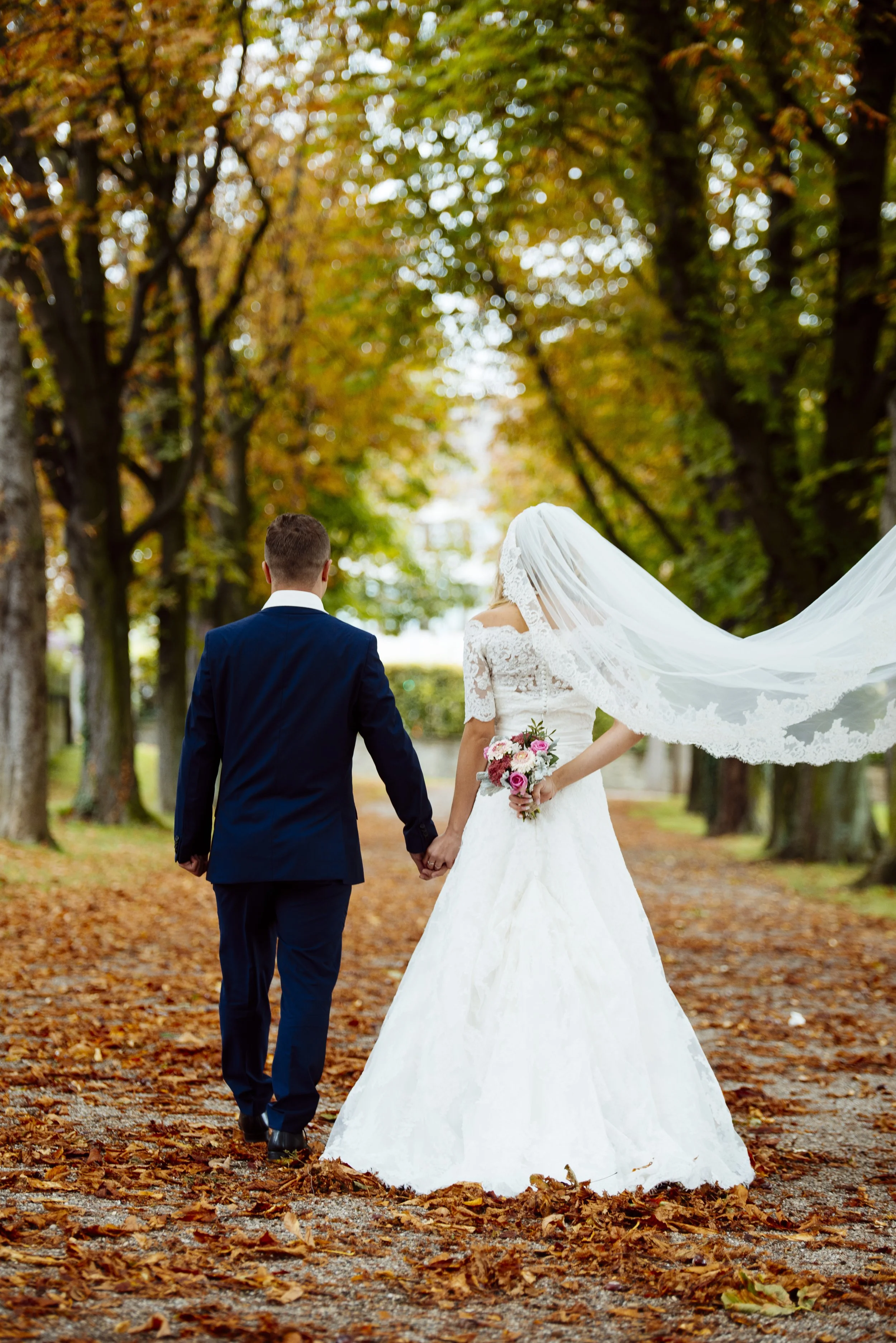 Ein Brautpaar, das Hand in Hand in einem Park im Herbst spazieren geht, die Blätter auf dem Boden sind orange und braun, die Bäume tragen herbstliche Farben.