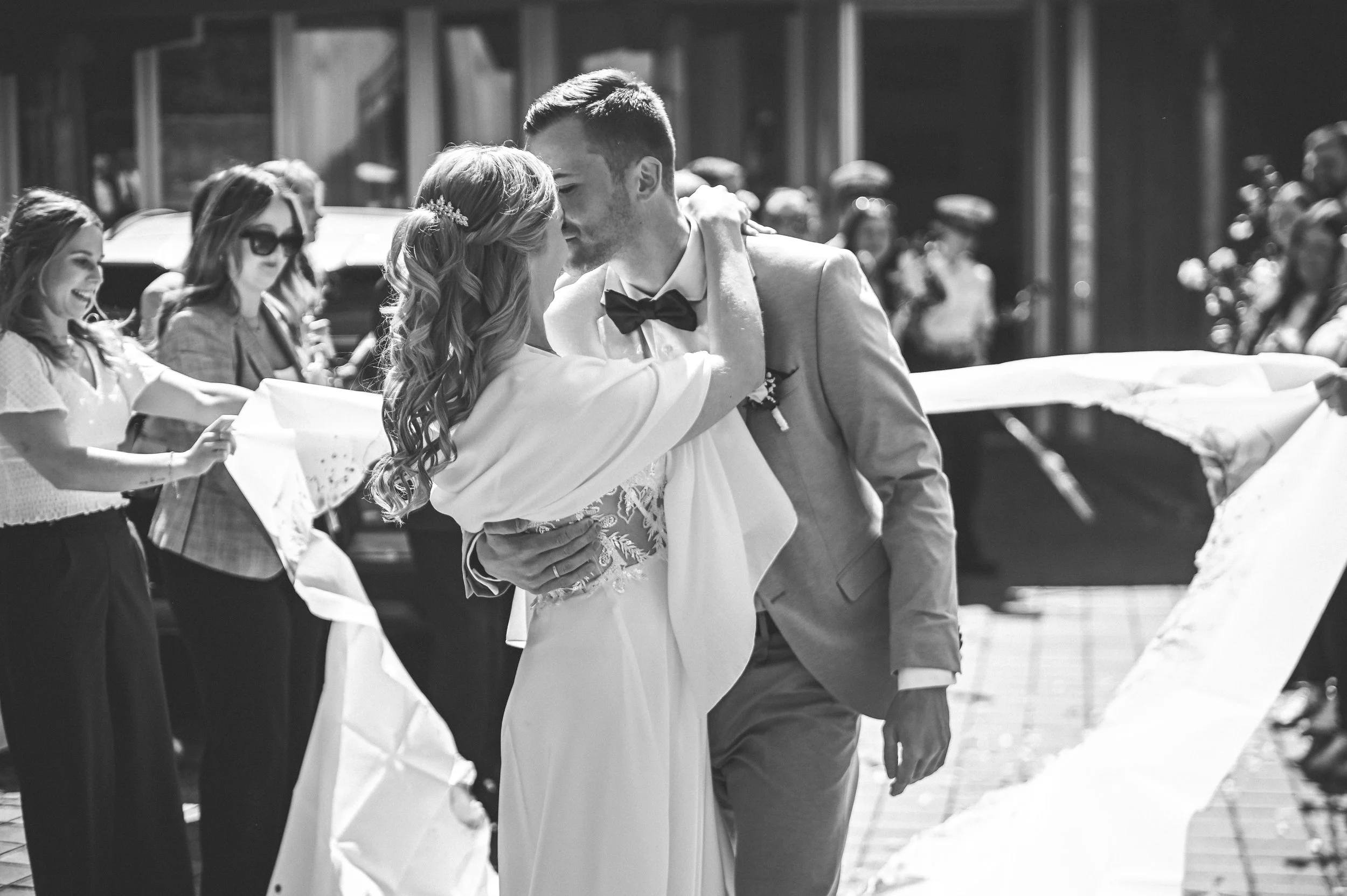 Paar bei einer Hochzeit, sich küssend, umgeben von Freunden und Familie, in einer lebhaften festlichen Atmosphäre.