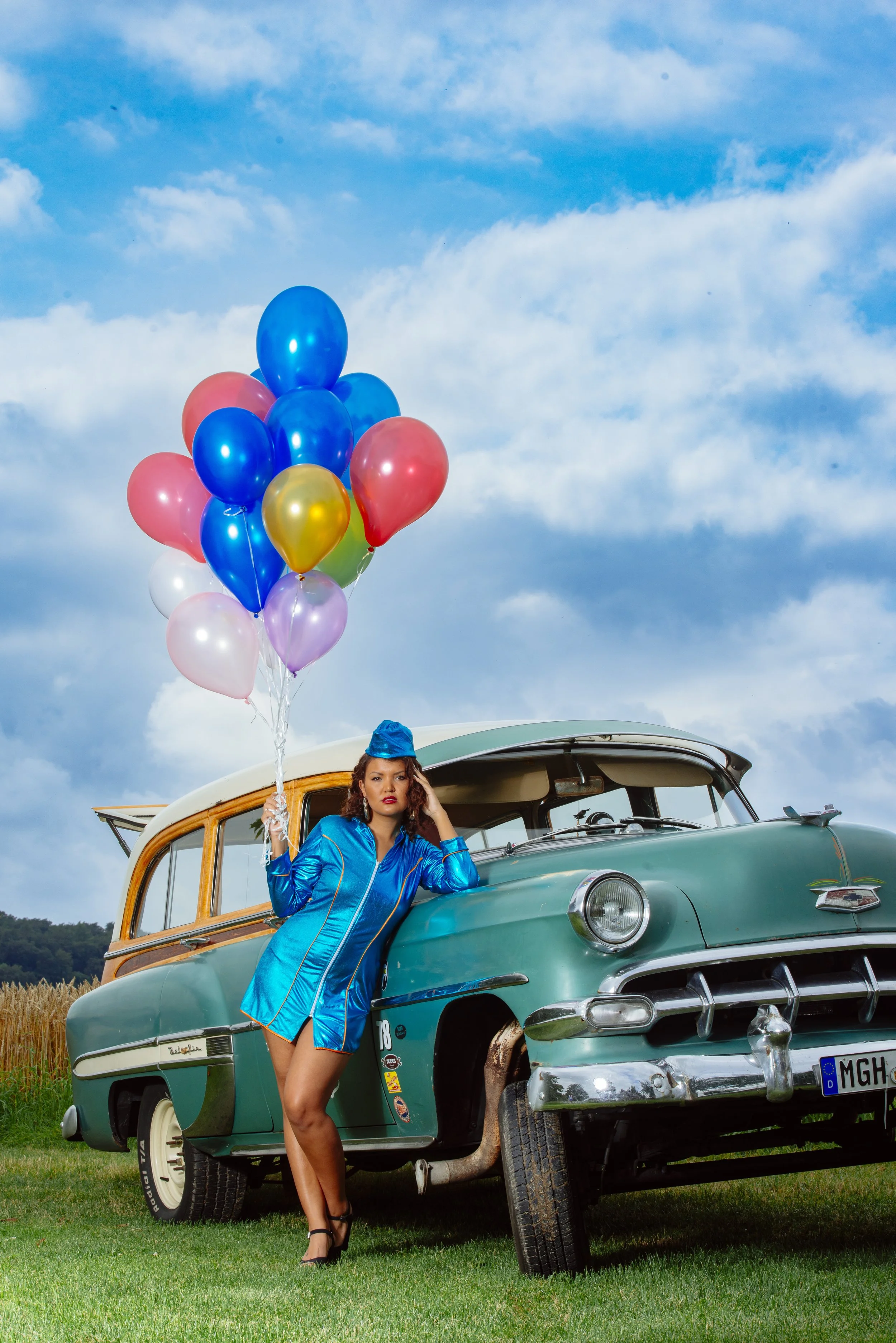 Eine Frau in einem blauen Anzug steht neben einem alten grünen Auto, hält eine bunte Ballonscheine und trägt einen blauen Hut. Das Auto ist in einer ländlichen Gegend mit Feldern im Hintergrund.