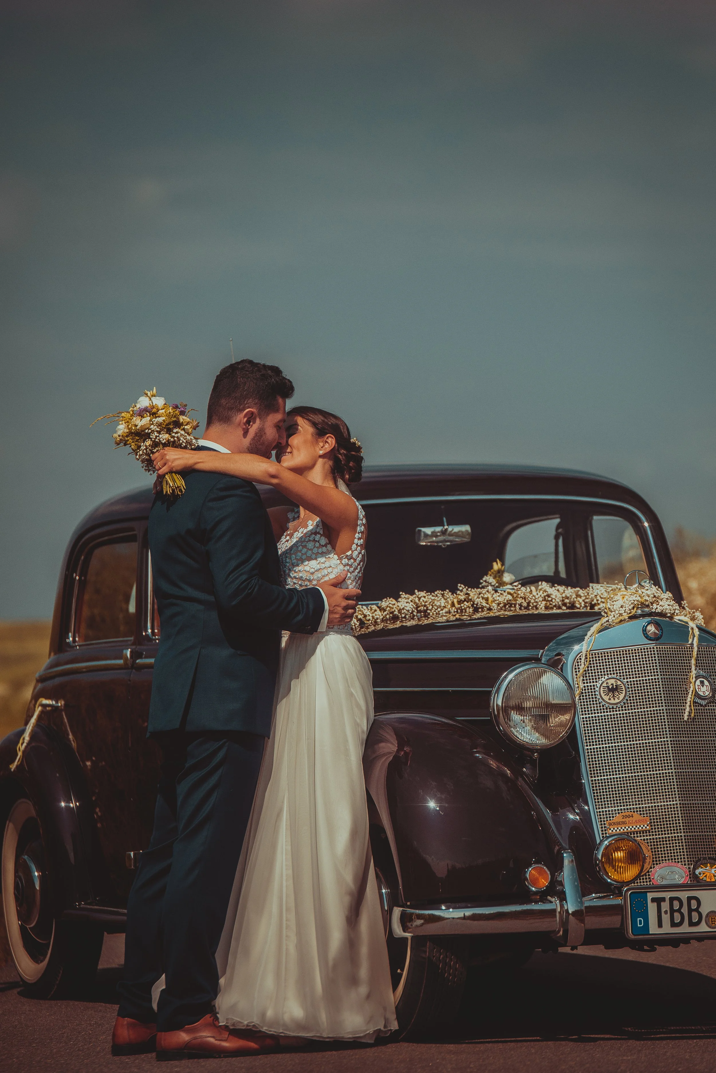 Ein Brautpaar steht vor einem schwarzen Oldtimer, der mit Blumen geschmückt ist. Das Paar umarmt sich und blickt sich liebevoll an, während die Frau ein weißes Brautkleid trägt und der Mann einen dunklen Anzug. Der Himmel ist klar und blau.