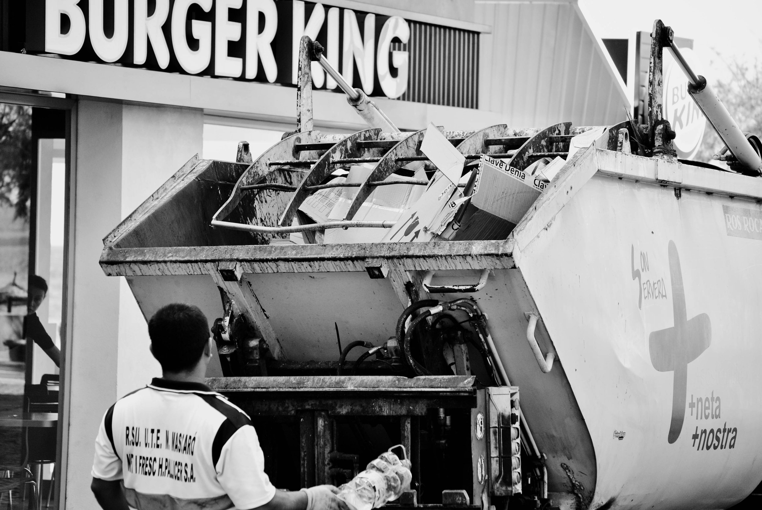 Ein Müllauto wird entladen, es sind Menschen im Vordergrund zu sehen, eines trägt eine Jacke mit Text. Im Hintergrund ein Geschäft mit dem Schild 'Burger King'.