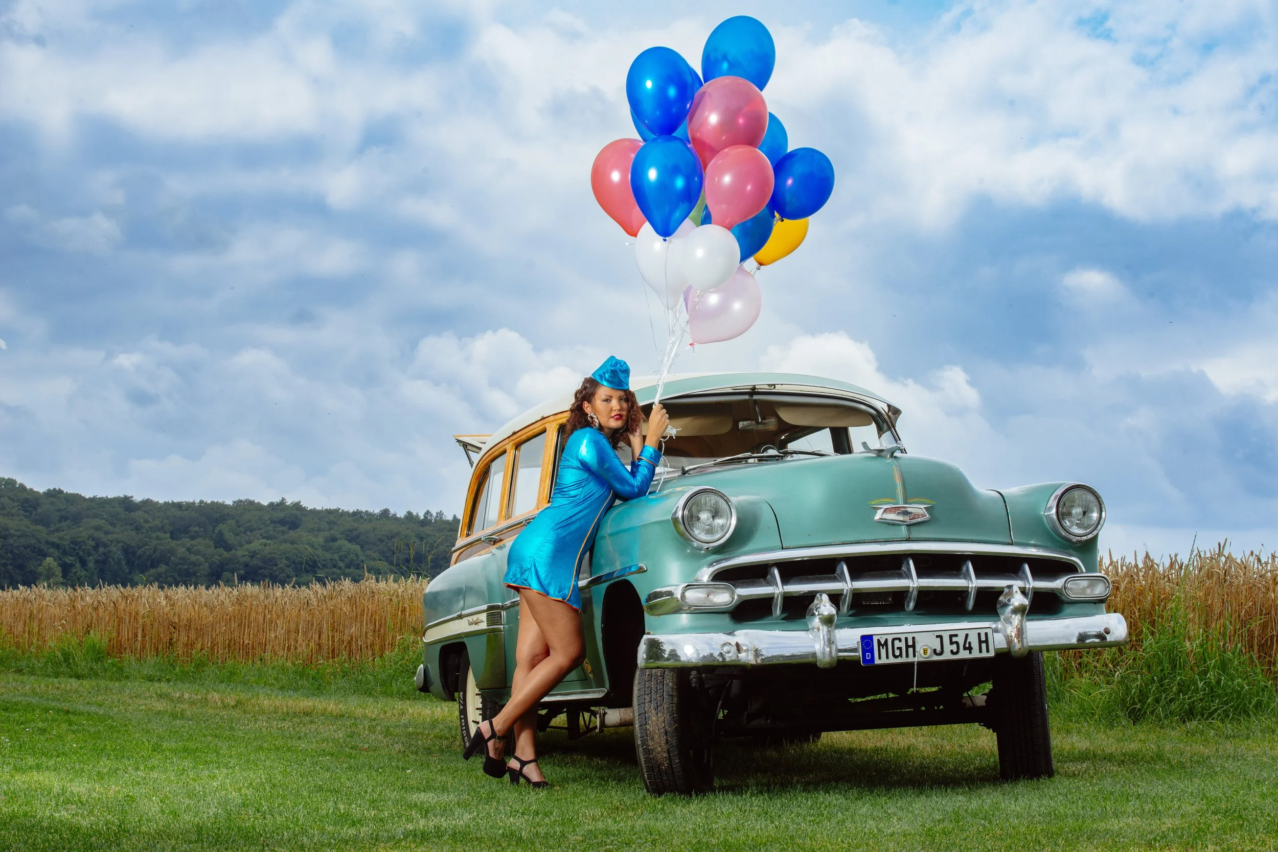 Eine Frau in einem blauen Kleid und schwarzen High Heels steht neben einem alten grünen Auto auf einem Feld. Sie hält einen Strauß bunter Ballons und trägt einen blauen Hut.