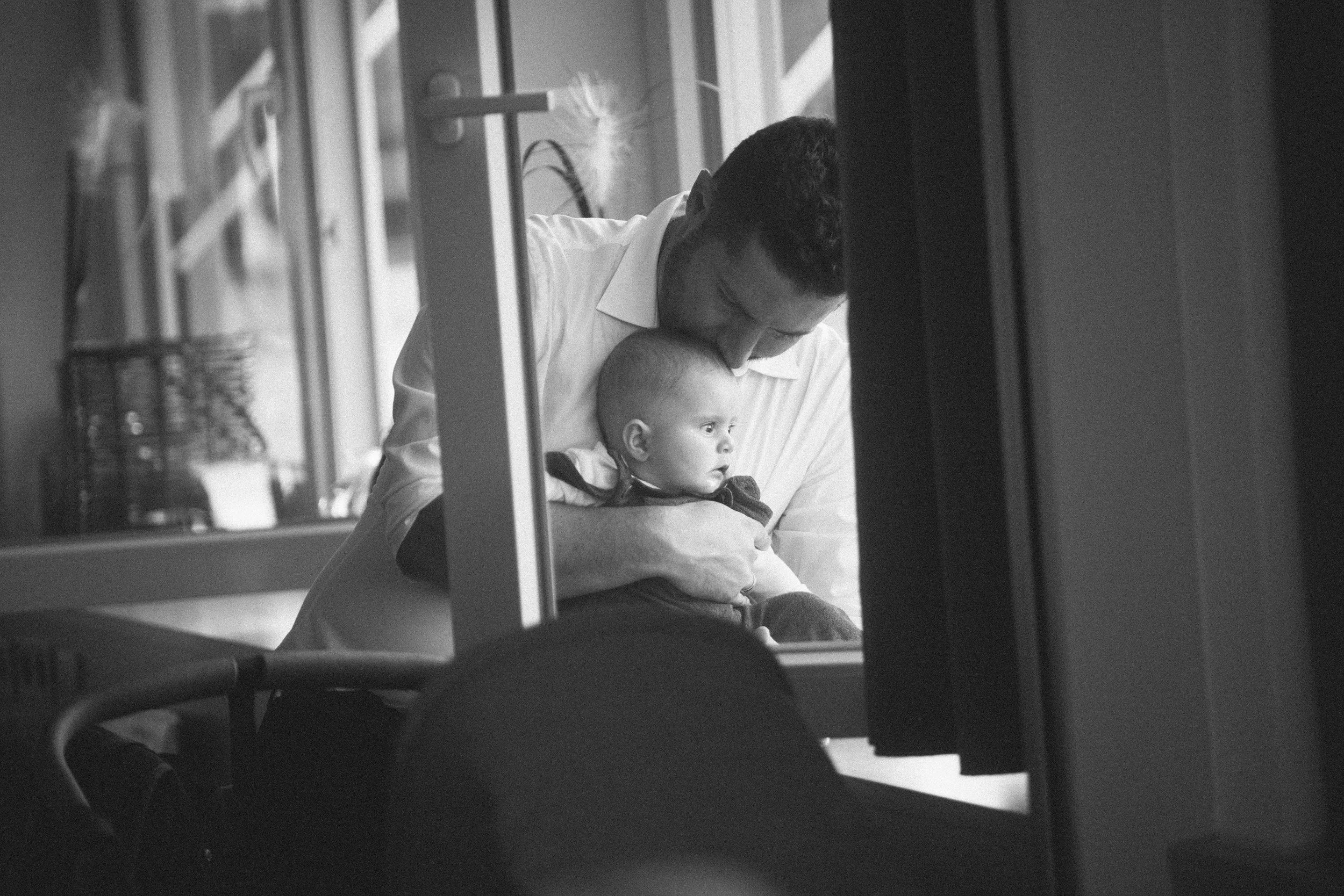 Ein Mann trägt ein neugeborenes Baby auf dem Arm und schaut durch ein Fenster nach draußen. Das Bild ist in Schwarz-Weiß.