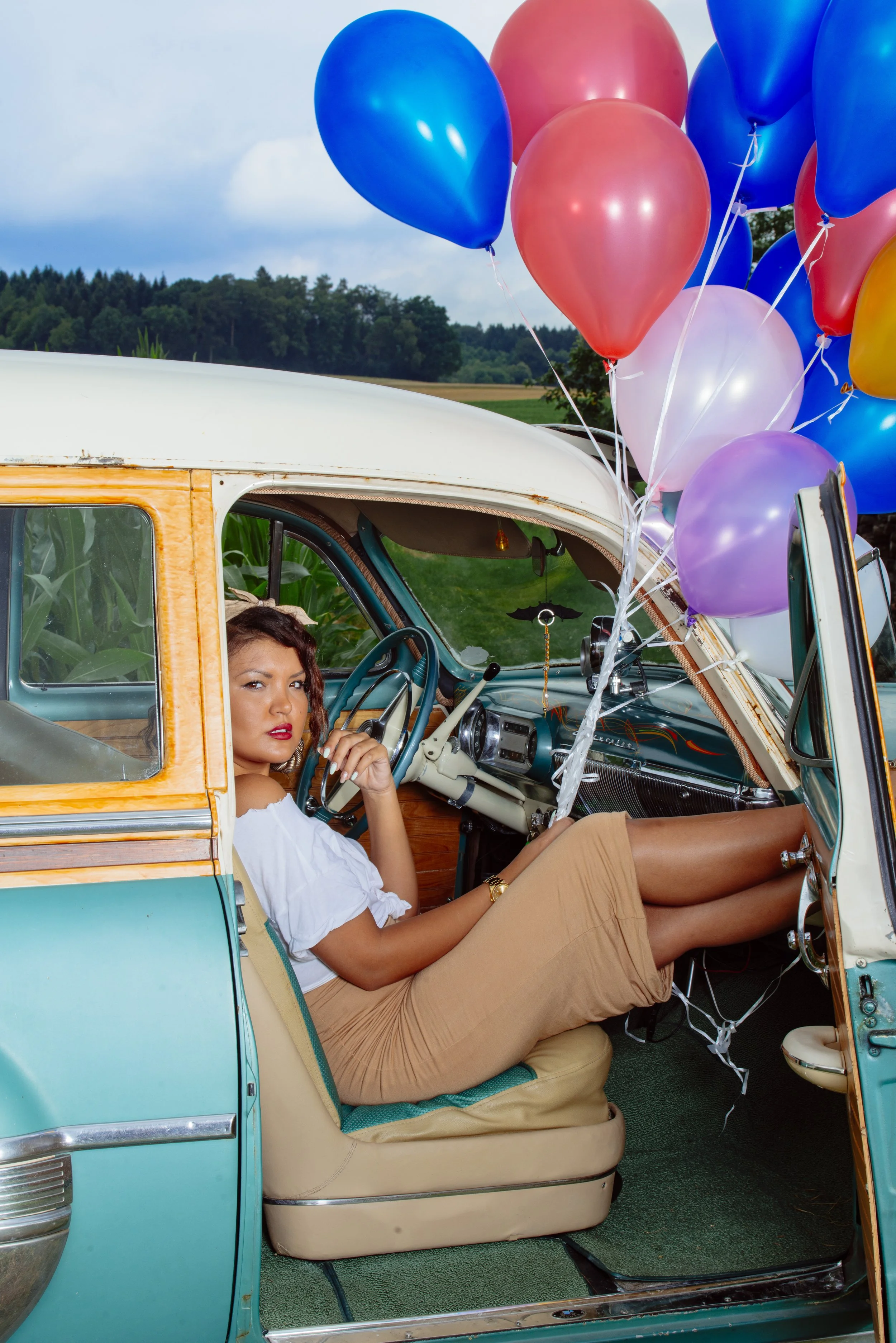 Junge Frau sitzt im Vintage-Auto mit bunten Ballons vor ländlicher Landschaft.
