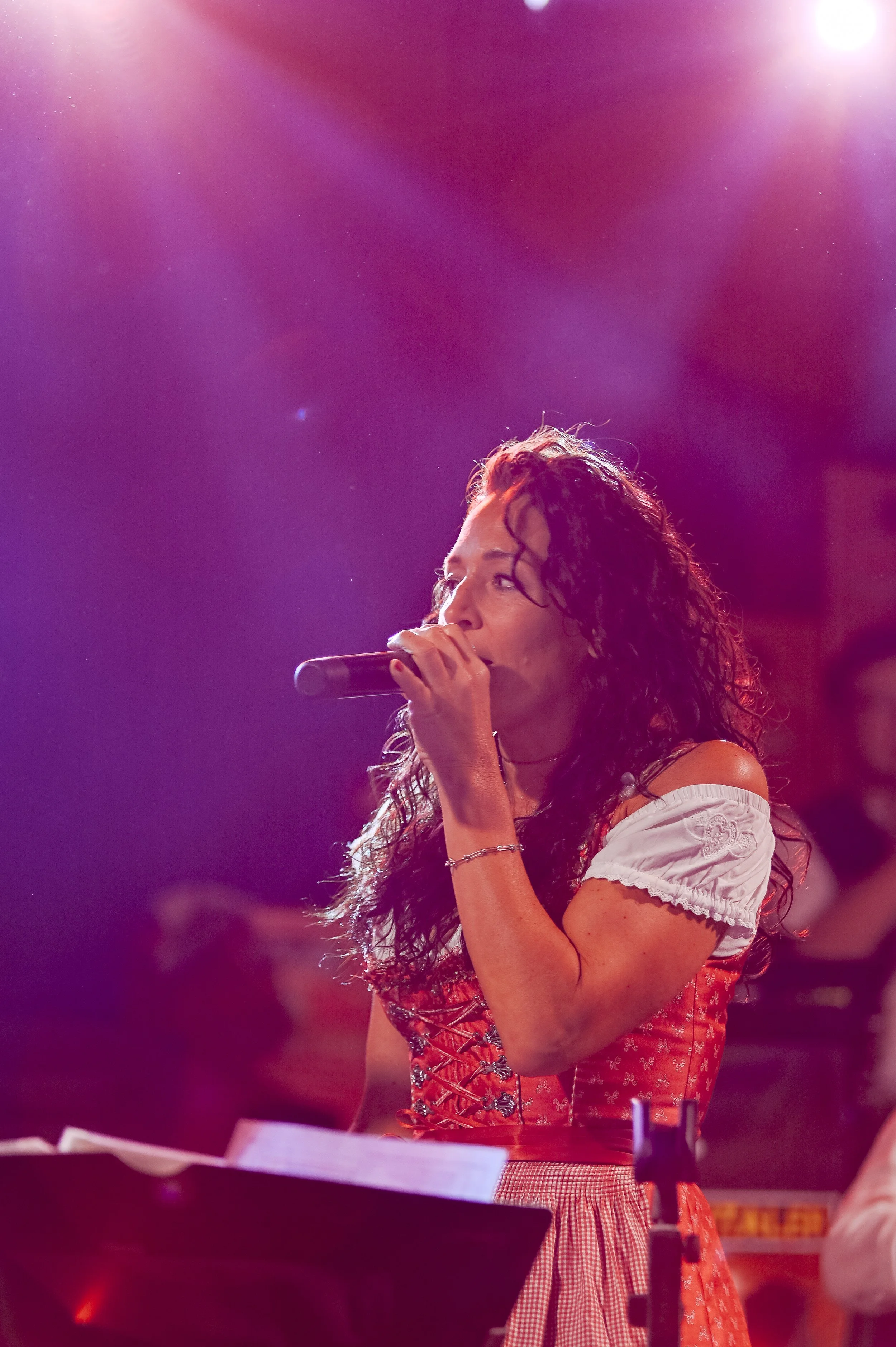 Junge Frau mit dunklen, lockigen Haaren singt mit einem Mikrofon bei einer Live-Performance, im Hintergrund bunte Lichter, vermutlich auf einer Bühne.