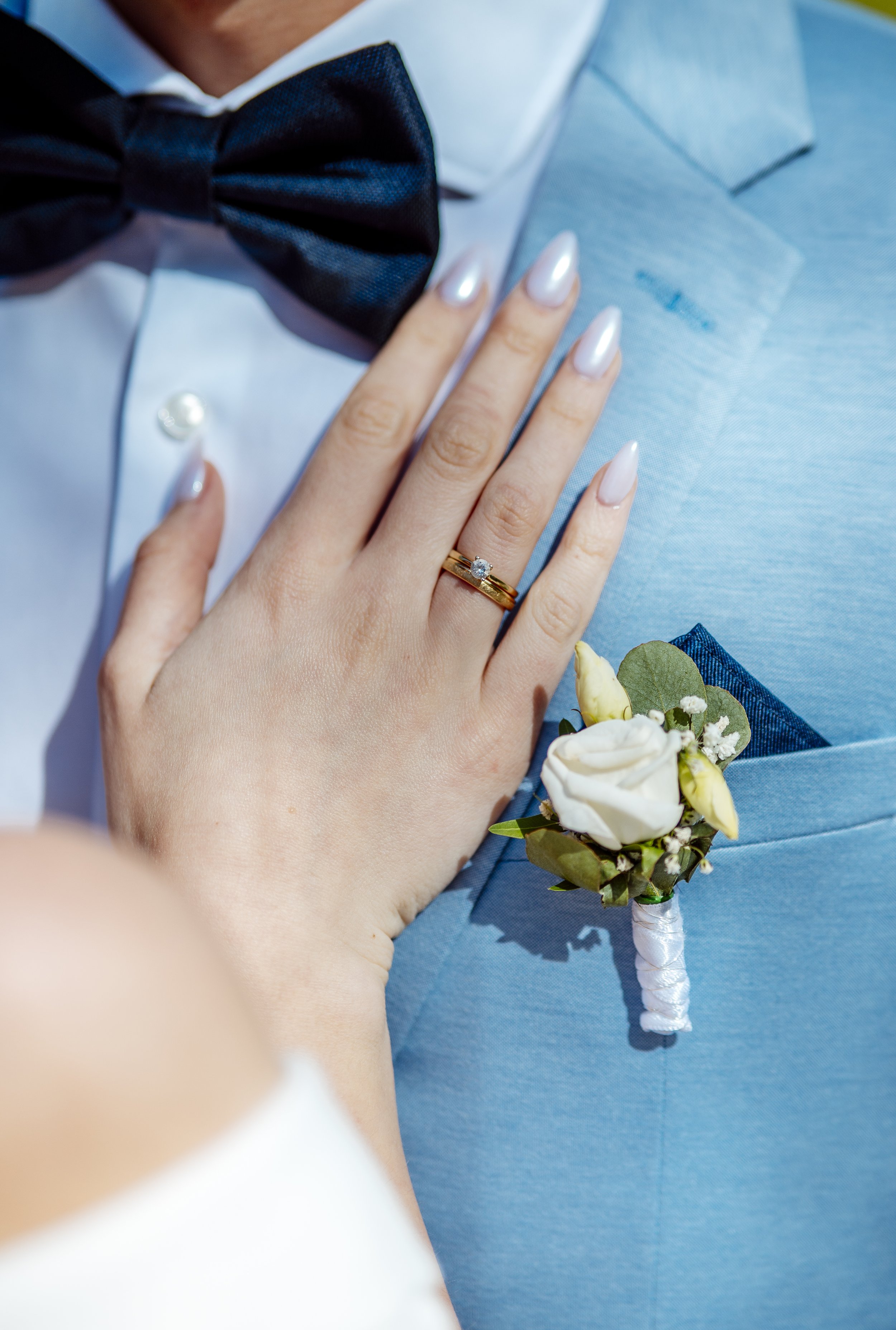 Nahaufnahme eines Paares bei ihrer Hochzeit, Fokus auf Hand mit Ehering auf der Brust des Bräutigams in blauer Jacke, daneben ein Blumenstrauß.