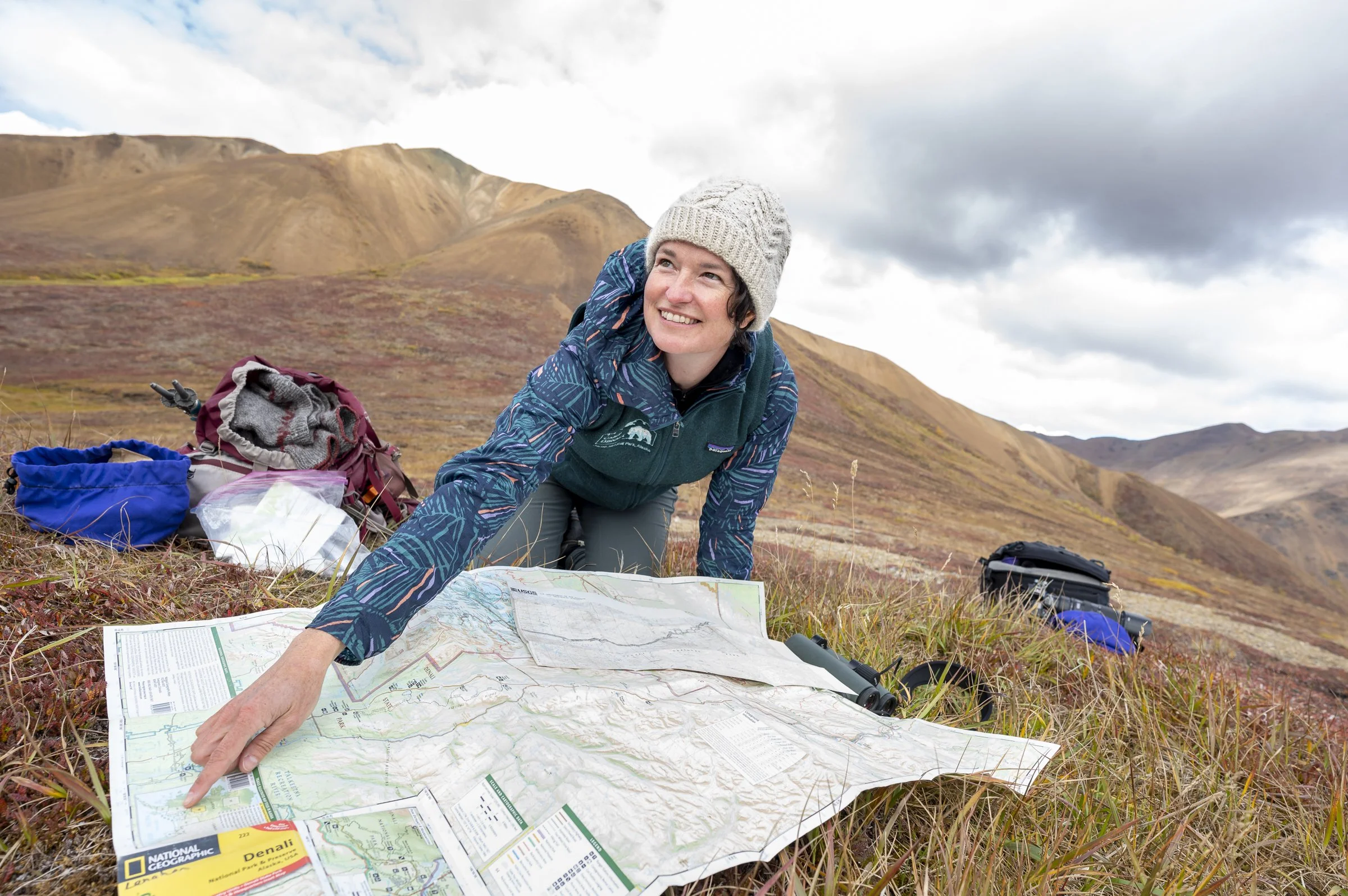 Woman in outdoor gear, smiling, wearing a knit cap, lying on a grassy hillside with a map and backpacks, with mountains and cloudy sky in the background.