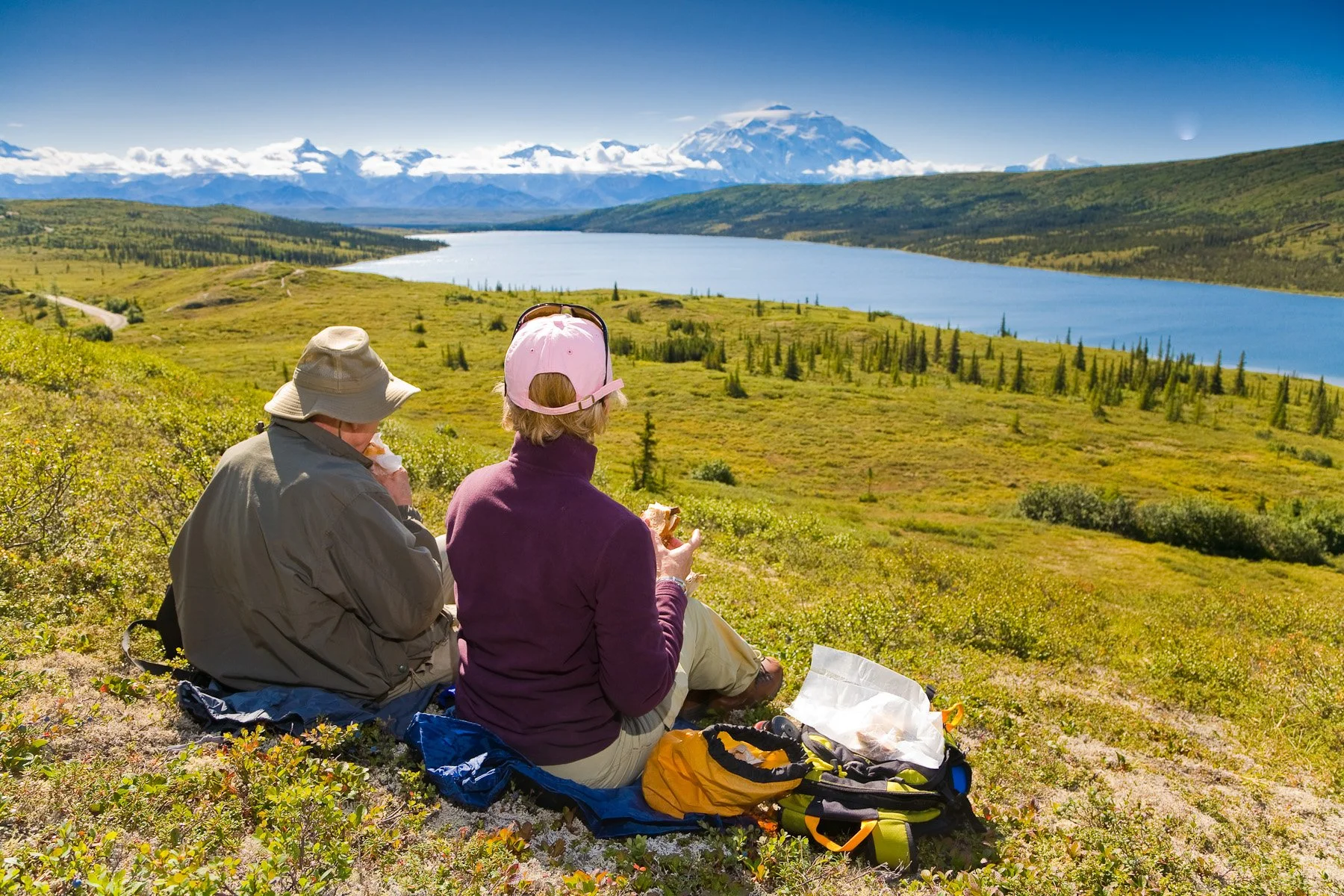 Two hikers sitting on a grassy hillside, overlooking a lake with mountains in the background, eating snacks. One wears a beige hat and jacket, the other a pink cap and purple jacket. Scenic landscape with snow-capped mountains and blue sky.