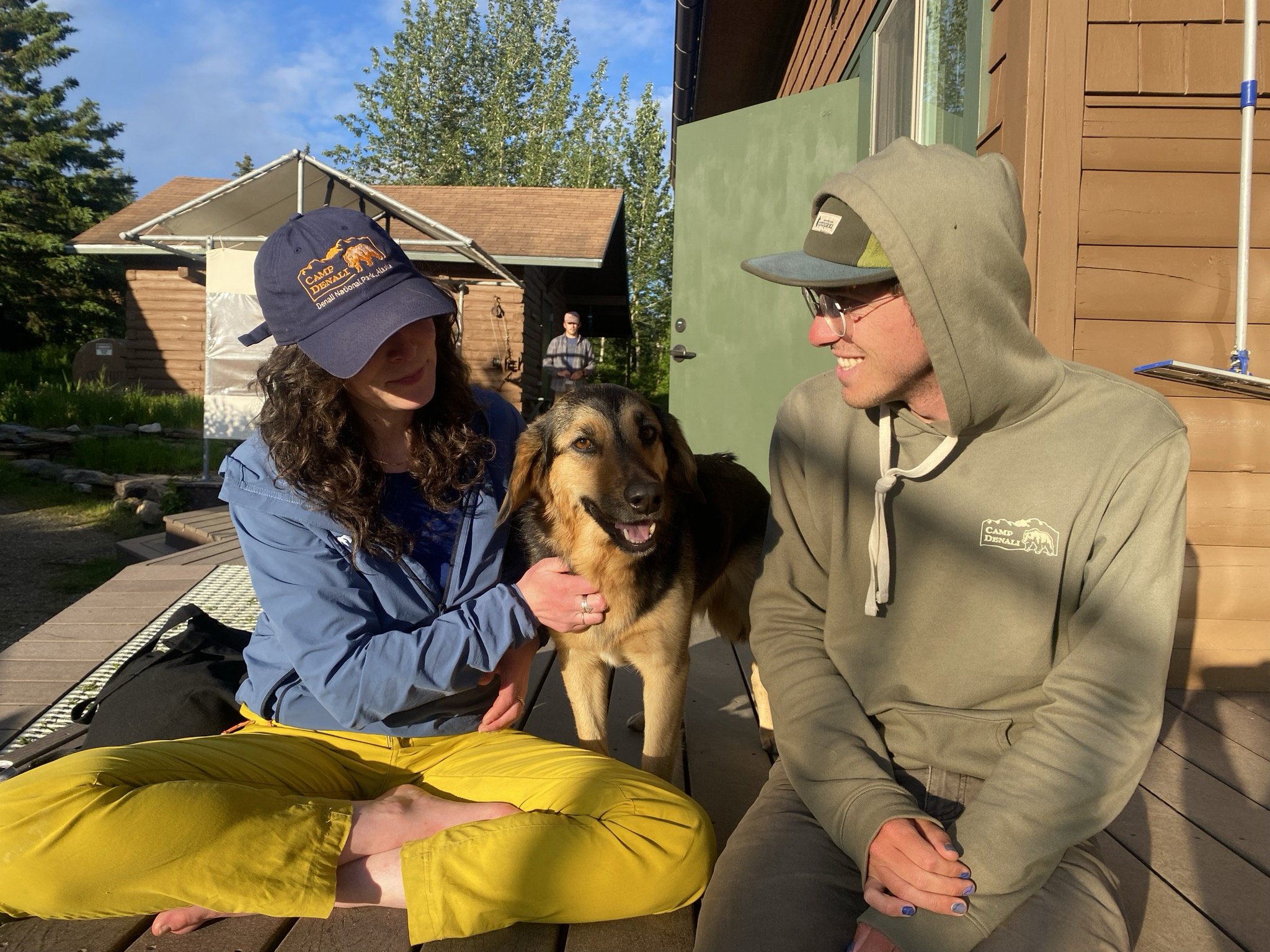 Two people, a woman in a blue jacket and a man in a green hoodie, are sitting outside on a wooden deck with a dog between them. The woman is wearing a navy cap with camp insignia and the man is wearing glasses and a gray cap. In the background, there is a small building, trees, and a clear blue sky.