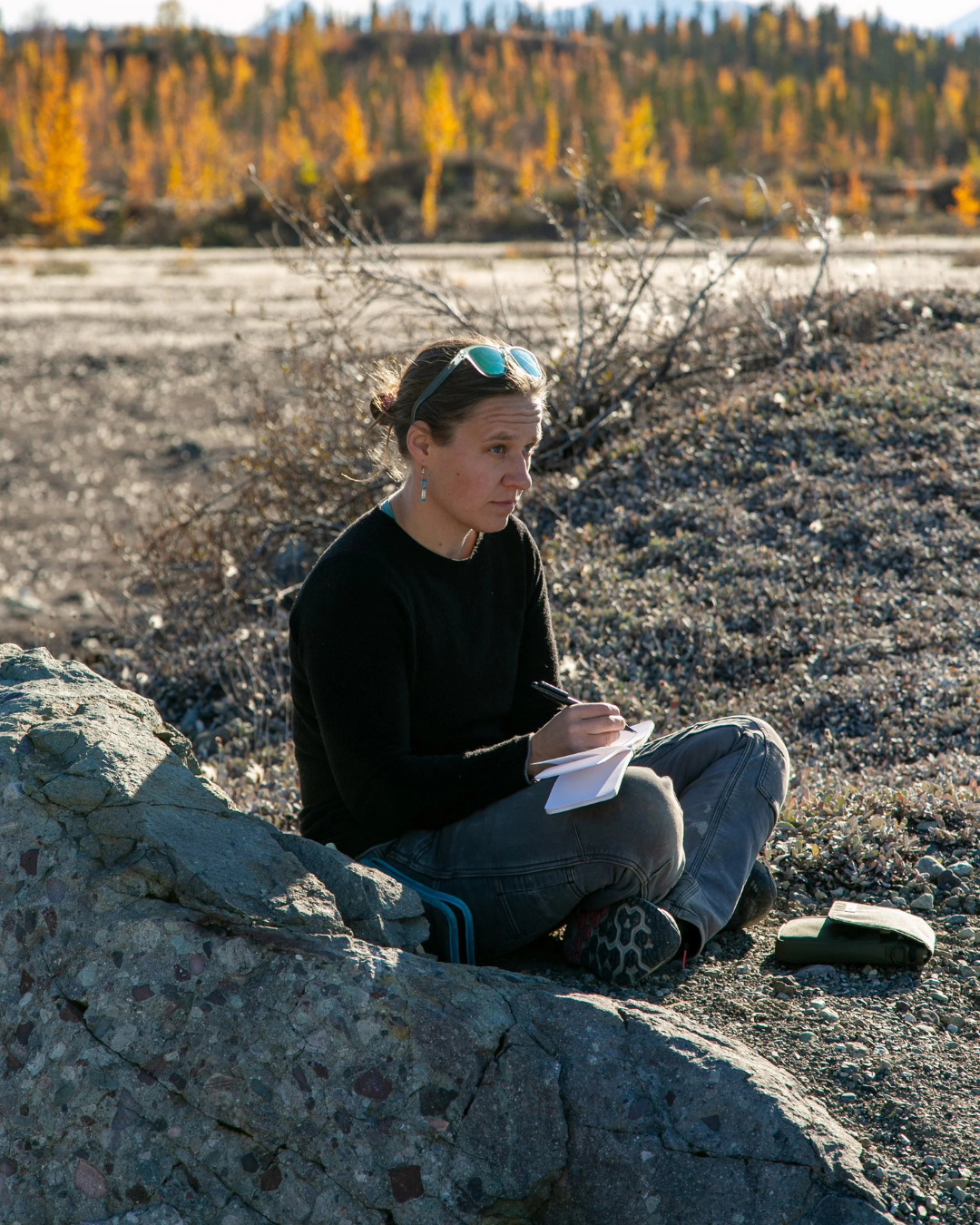 A woman sitting on a rock outdoors near a river, writing in a notebook with a pen, wearing a black shirt, gray pants, and sunglasses on her head, with a background of autumn trees and clear weather.