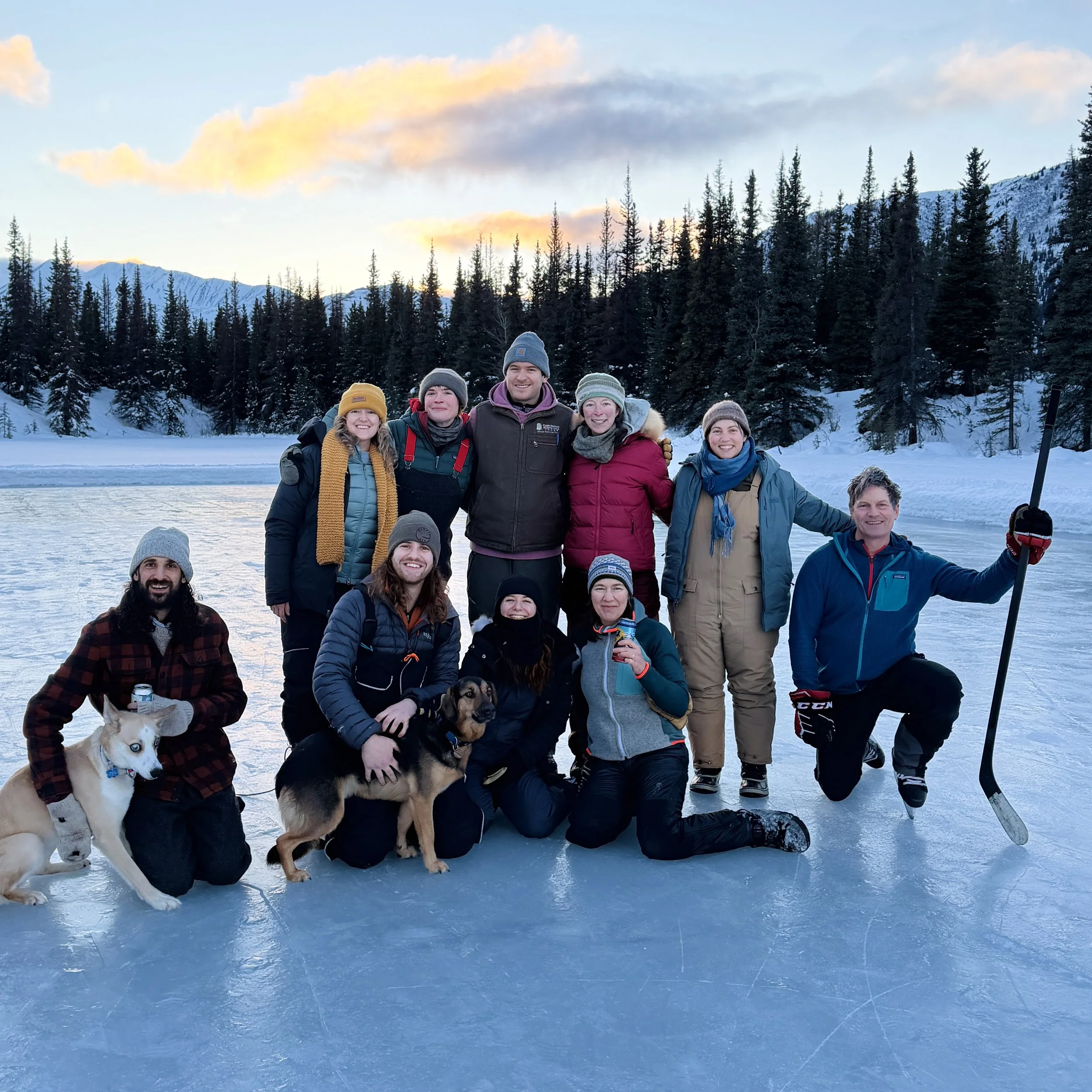 A group of people and dogs standing on a frozen lake with snow-covered mountains and pine trees in the background during sunset.