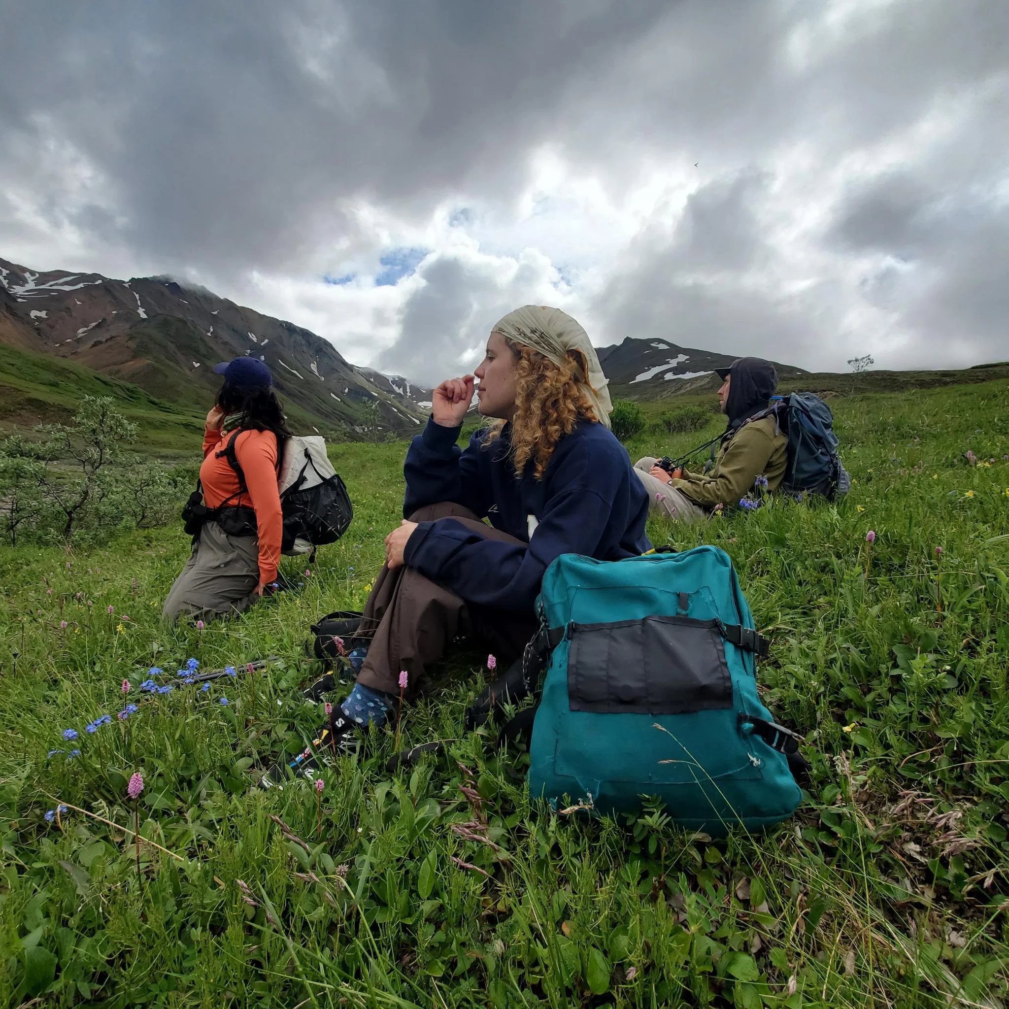 A group of hikers sitting on a grassy field with mountains and a cloudy sky in the background. The hikers are wearing outdoor clothing and backpacks.