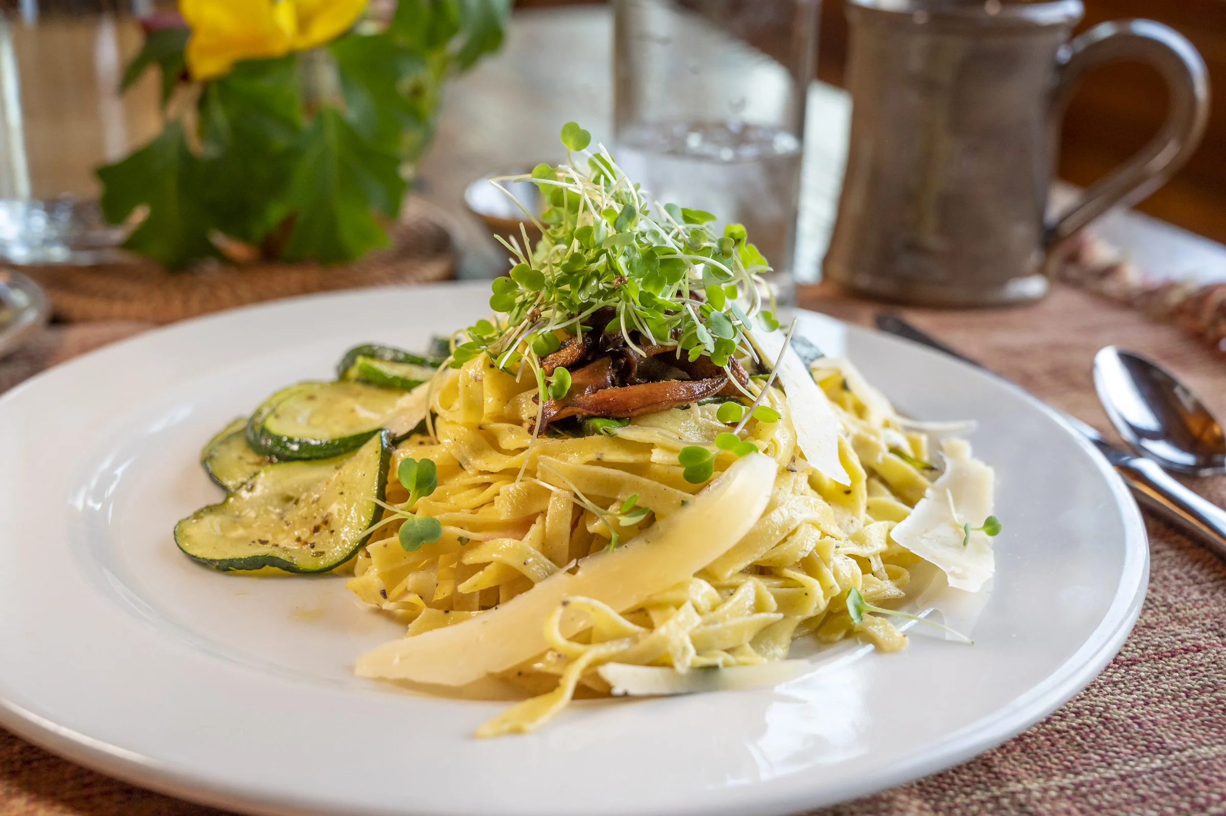 Plate of fettuccine pasta with zucchini slices and microgreens, topped with mushrooms and Parmesan cheese. In the background, there is a plant, a glass of water, and a brown mug on a wooden table.