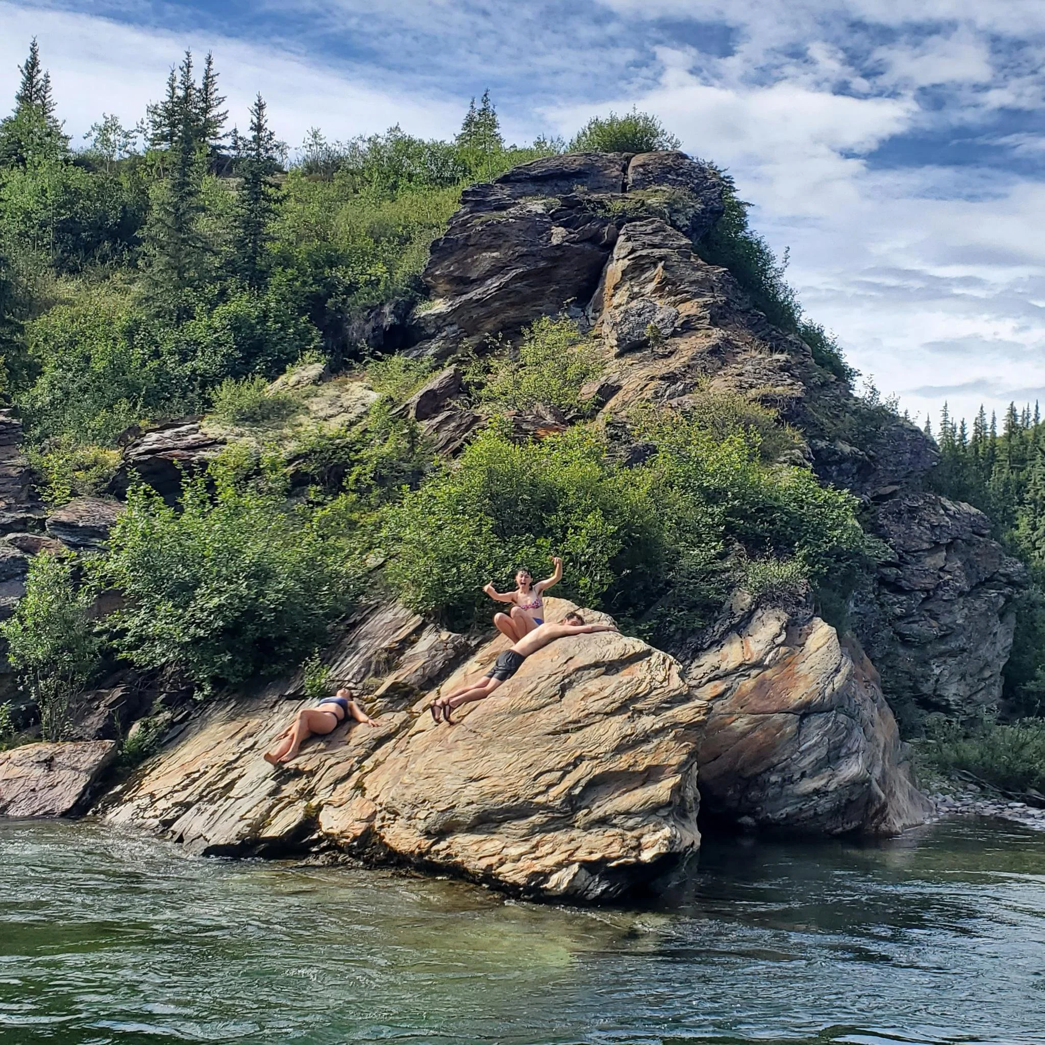 A rocky cliff with greenery at the base and trees at the top, with a person sitting on the rocks near the water at the bottom of the cliff.