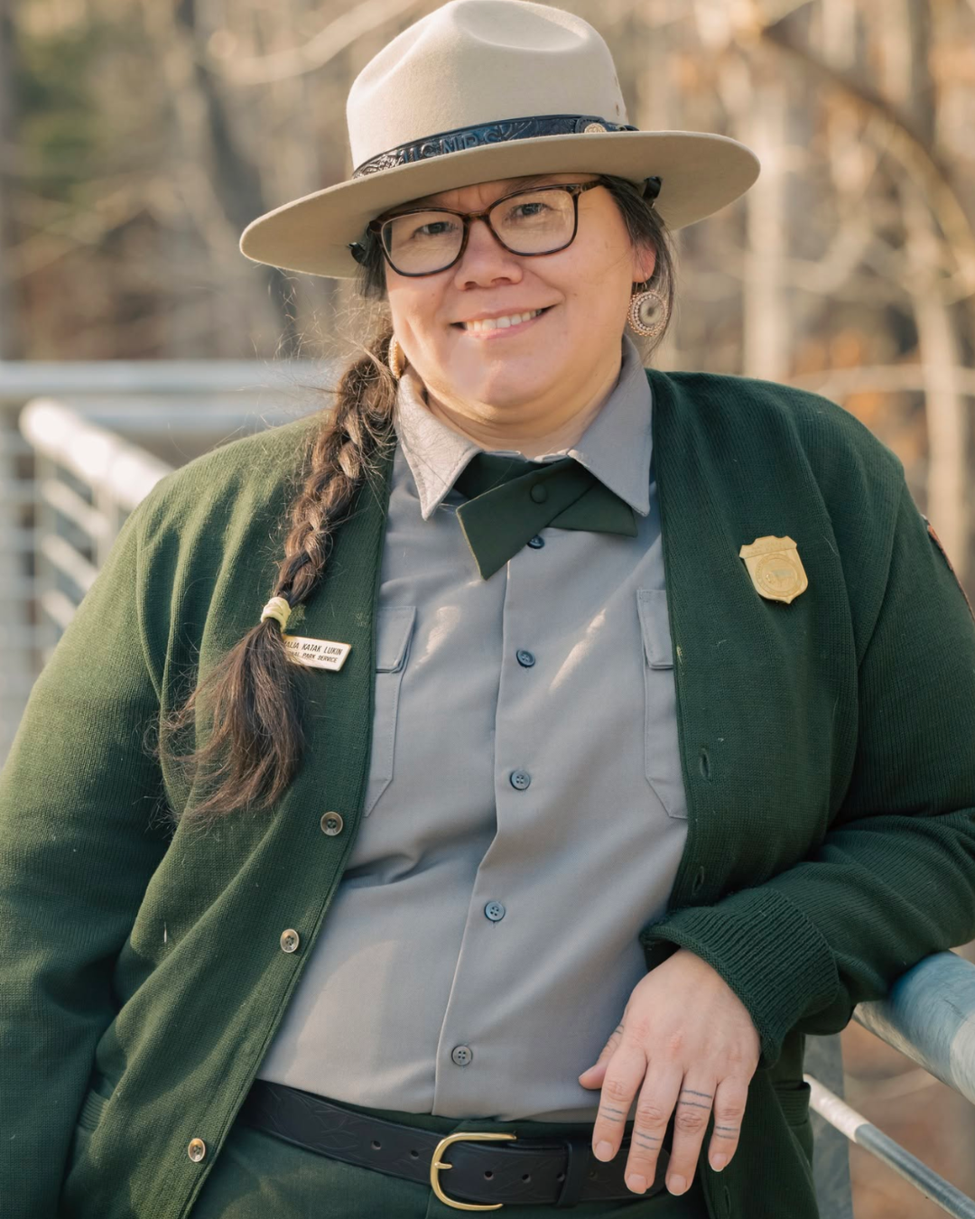 A woman smiling outdoors, wearing a ranger uniform with a badge, a name tag, a green jacket, a wide-brimmed beige hat, and glasses.