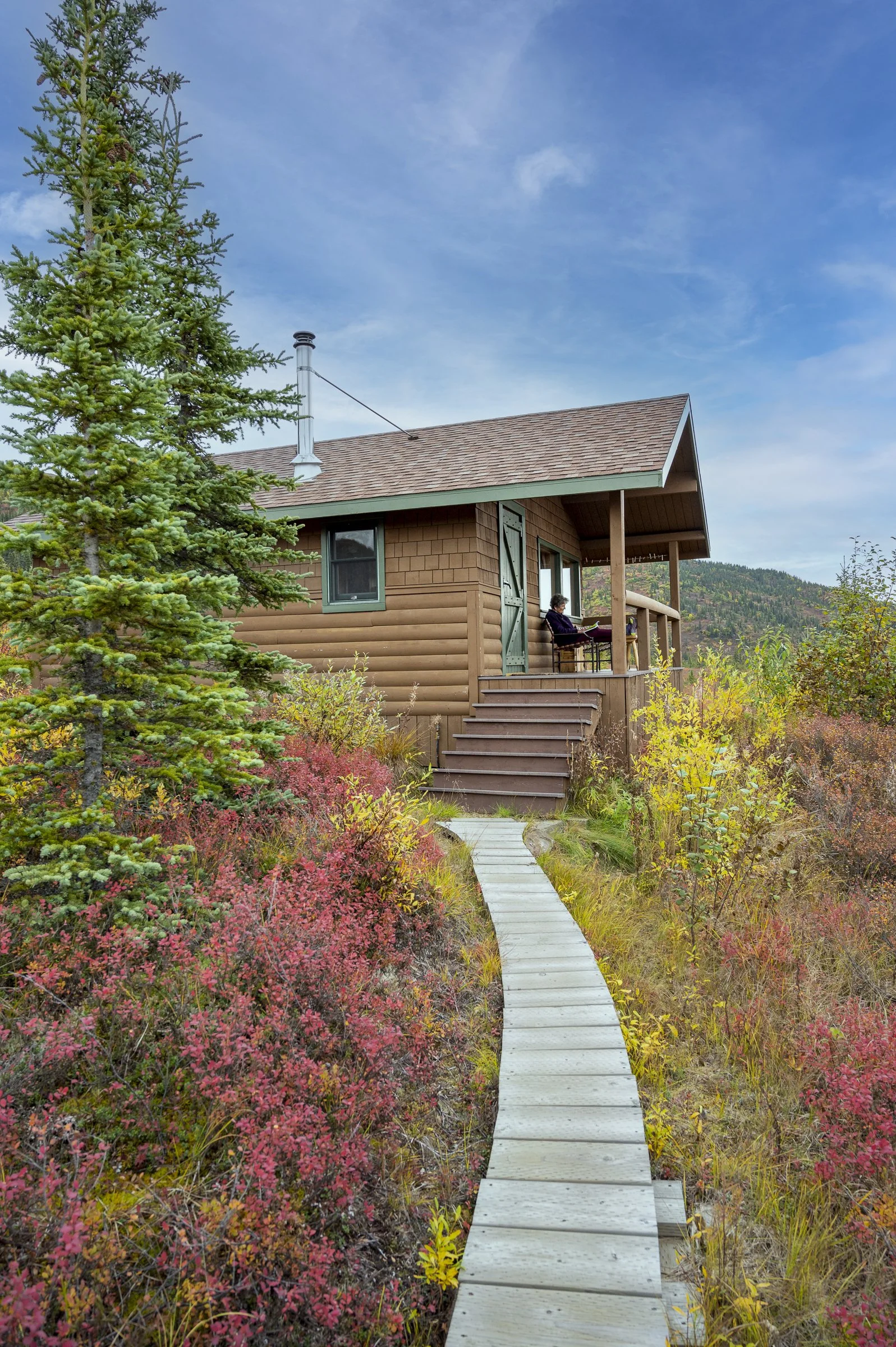 A wooden cabin with a green door and window, surrounded by colorful autumn foliage and a pathway leading to it. A person sits on the porch, reading.