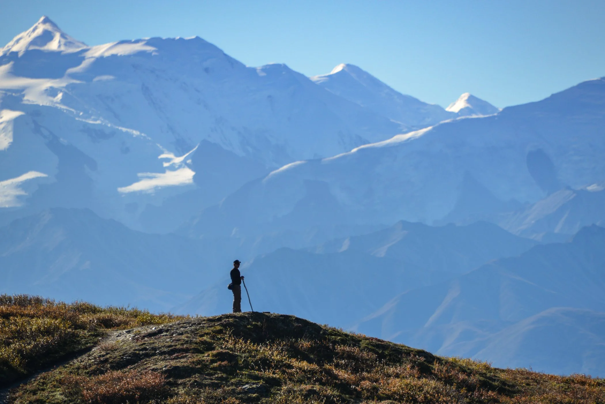 A person standing on a grassy hilltop with a walking stick against a backdrop of snow-capped mountains and a clear blue sky.