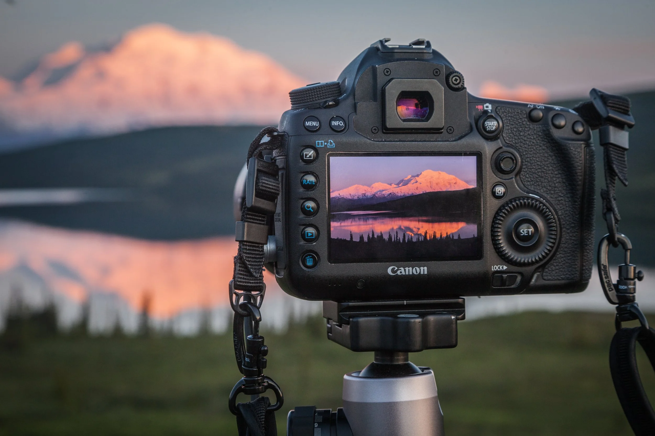 A Canon camera on a tripod captures a snapshot of a mountain during sunset, with the mountain side illuminated in a pinkish glow.
