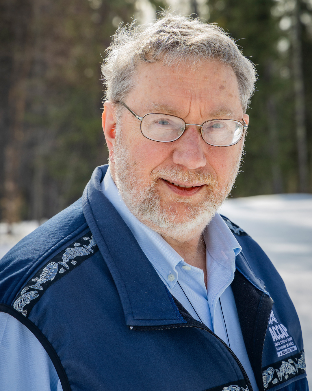 A smiling elderly man with glasses, gray hair, and a beard, wearing a blue vest over a light blue shirt, outdoors with trees in the background.