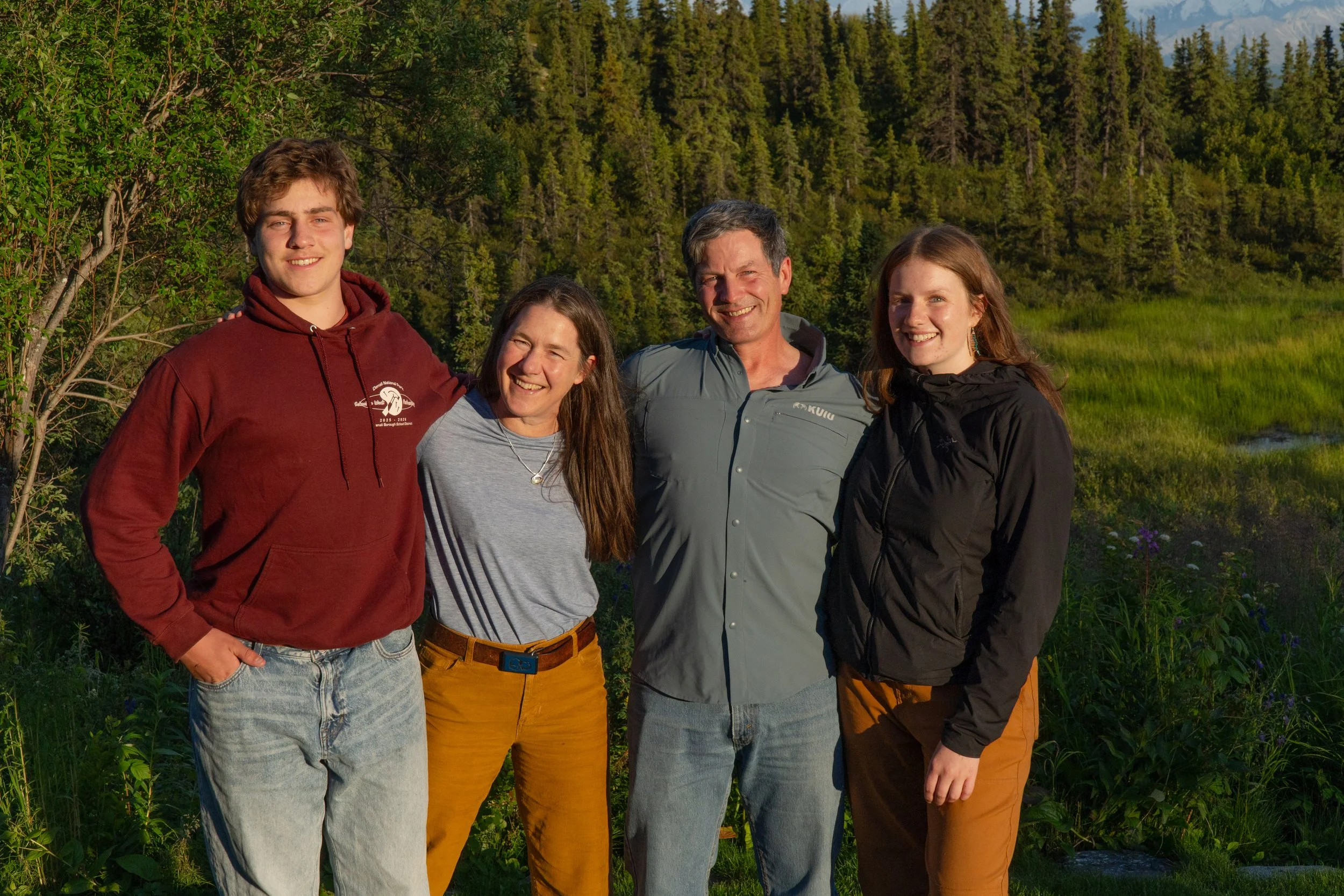 A group of five smiling people standing outdoors in a lush green natural setting with trees and grass, enjoying a sunny day.