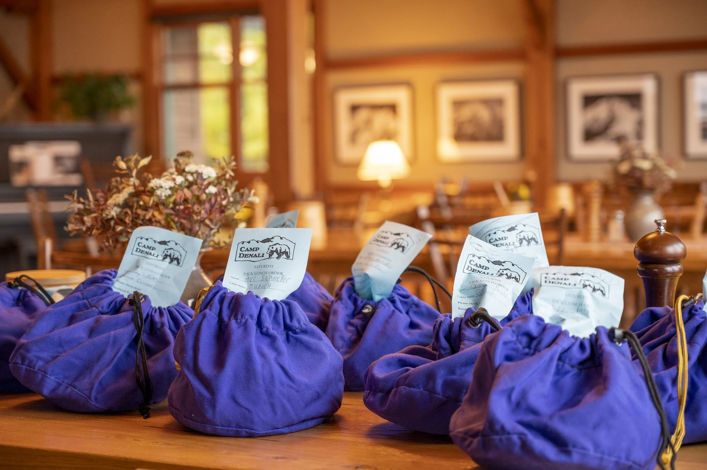 A table with purple drawstring backpacks containing conference or event papers with the text "Camp Denali" and a mountain logo, inside a cozy wooden room with framed pictures on the wall, a lamp, and decorative flowers.