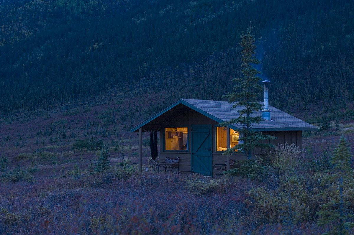 A small cabin in a forested area at dusk with warm yellow light glowing from the windows, surrounded by tall trees and low vegetation.