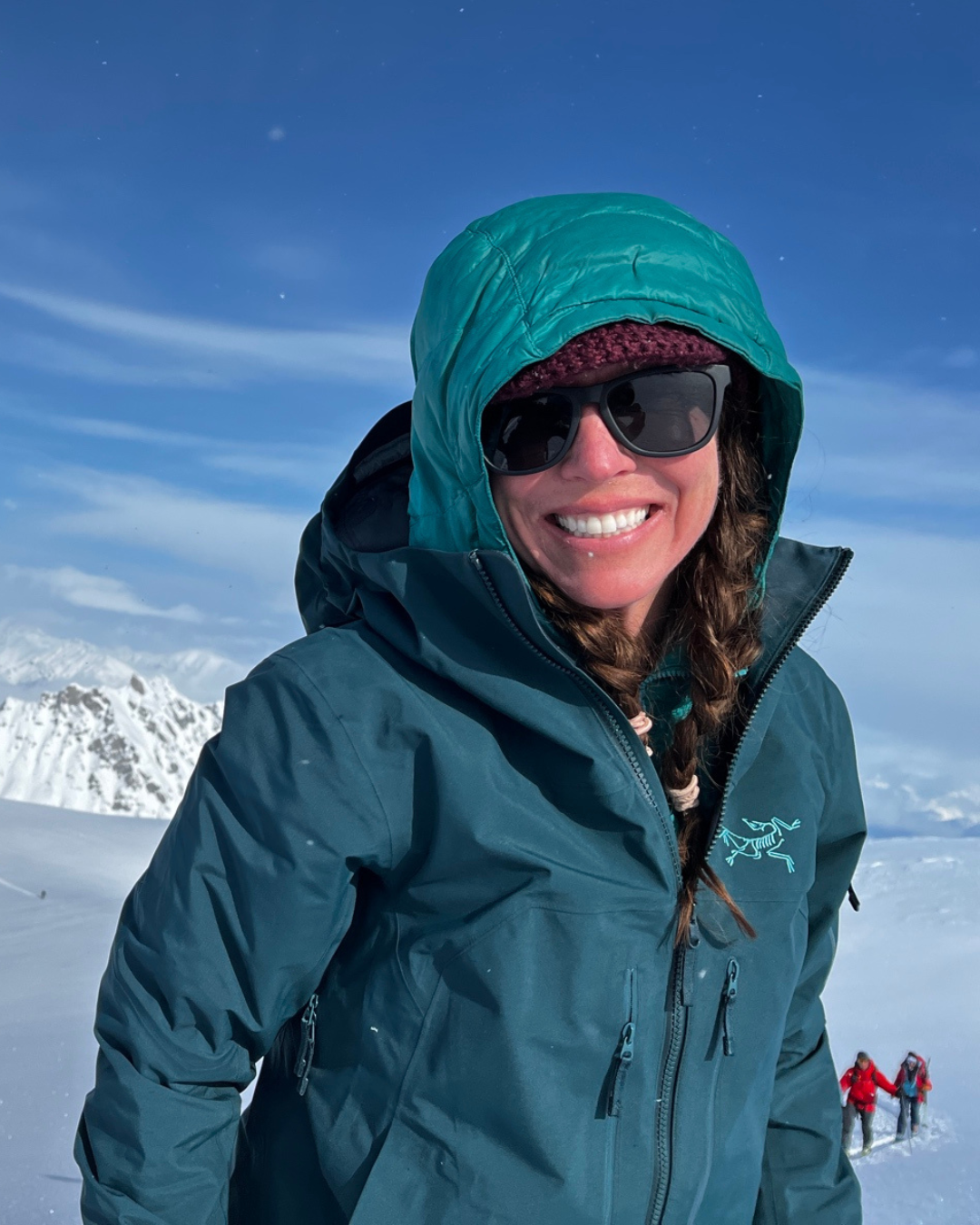A woman in a teal winter jacket, wearing sunglasses and a beanie, smiling in a snowy mountain landscape with a clear blue sky. In the background, two people are trekking in the snow.
