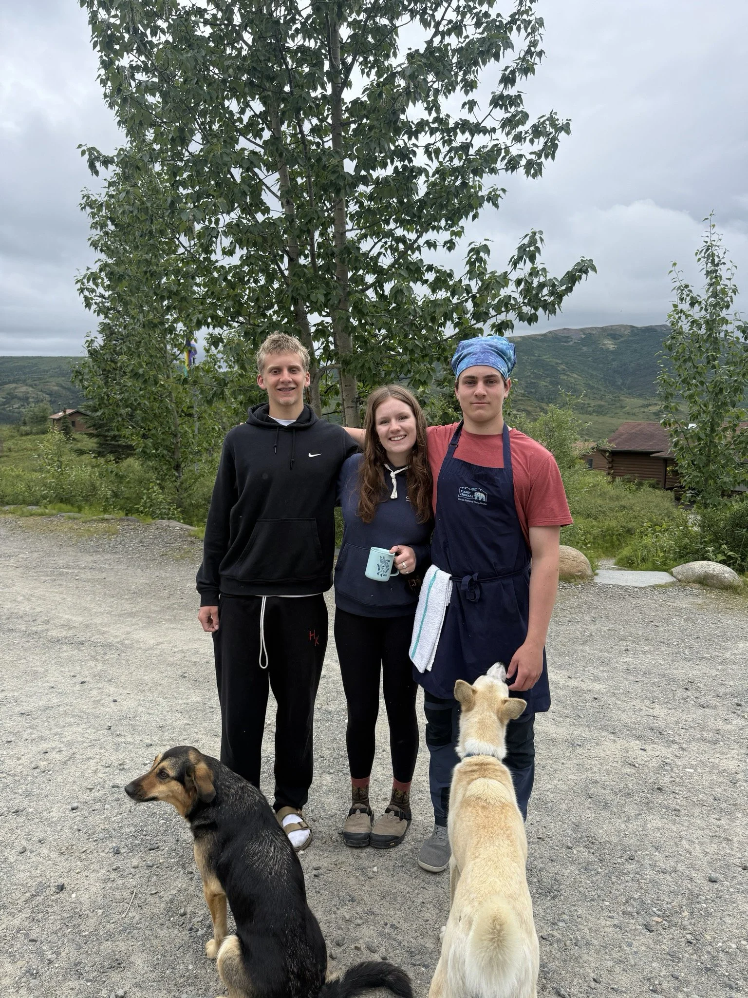 Three young people standing outdoors with two dogs, in a rural area with mountains and trees in the background, overcast sky.