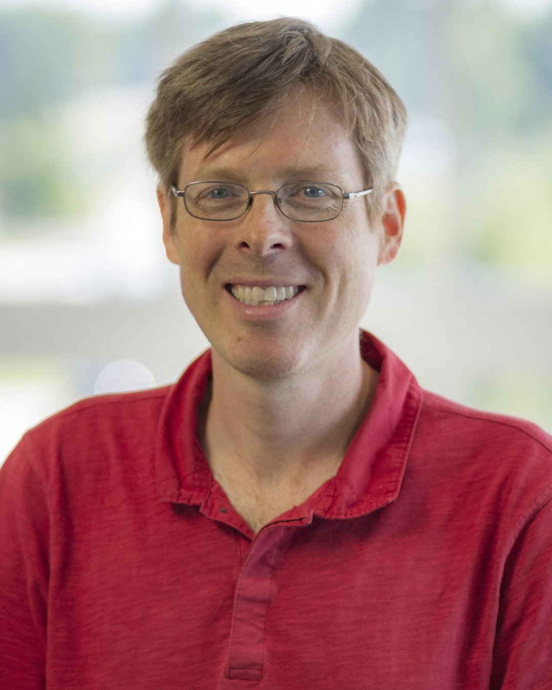 A smiling man with glasses, wearing a red polo shirt, in front of a blurred outdoor background.