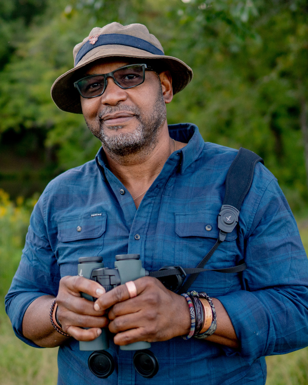 A man outdoors wearing a bucket hat, sunglasses, a blue shirt, and holding binoculars, with a backpack strap over his shoulder.