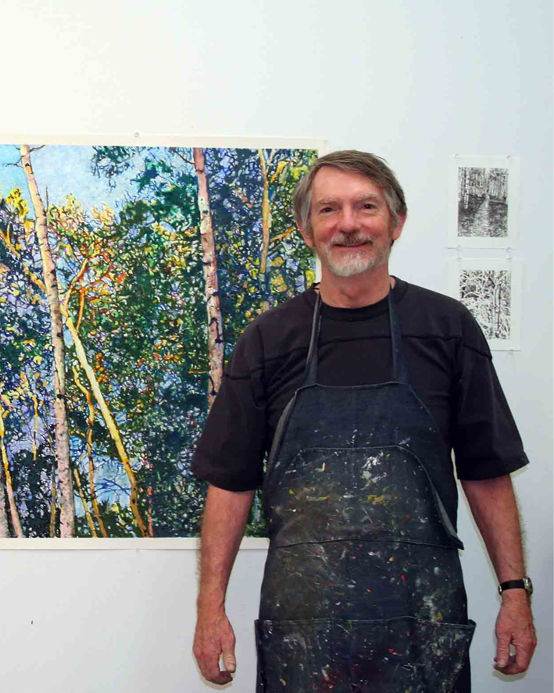 Man with gray hair and beard wearing a black shirt and apron standing in front of a colorful forest painting.