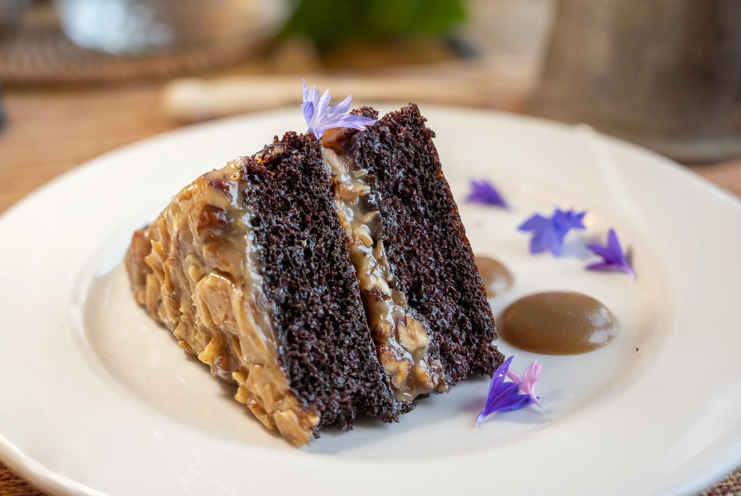 A slice of chocolate cake with caramel and nuts topping, garnished with purple edible flowers on a white plate