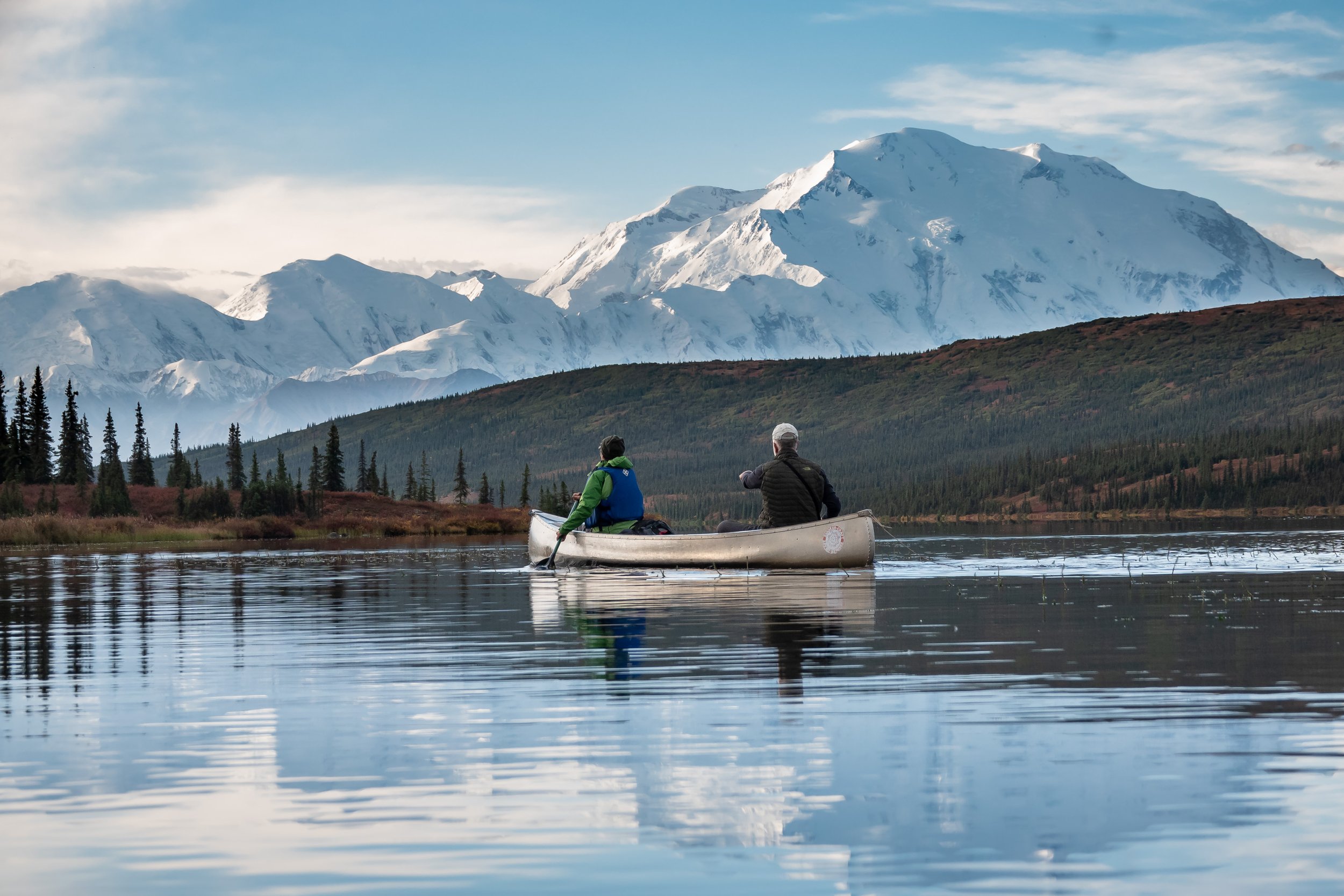 Two people in a canoe on a calm lake with snow-covered mountains in the background and a partly cloudy sky.