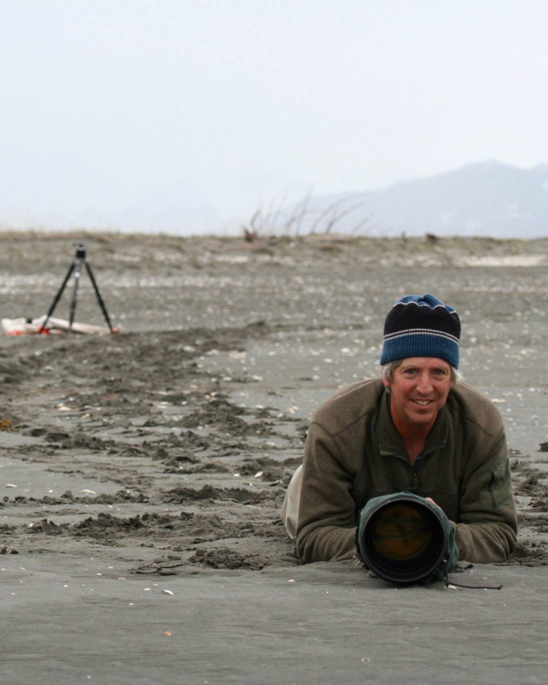 A man lying on a beach with a camera lens in front of him, wearing a beanie and a jacket, with a tripod set up in the background on the sand.