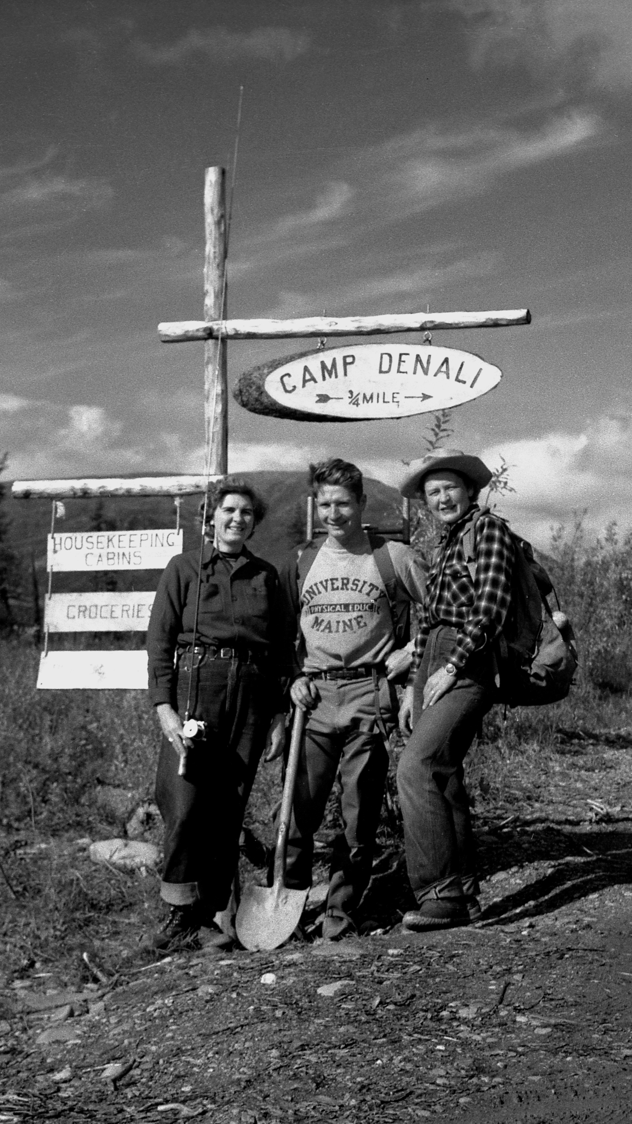 Three people hiking outdoors in front of a sign that reads 'Camp Denali, ¾ mile'. The group includes two women and one man, all smiling, wearing outdoor clothing, and carrying backpacks. The woman on the right wears a plaid shirt and a wide-brimmed hat. The man holds a shovel. The sign is attached to a wooden post with another sign below indicating 'Housekeeping Cabins' and 'Grocery'.