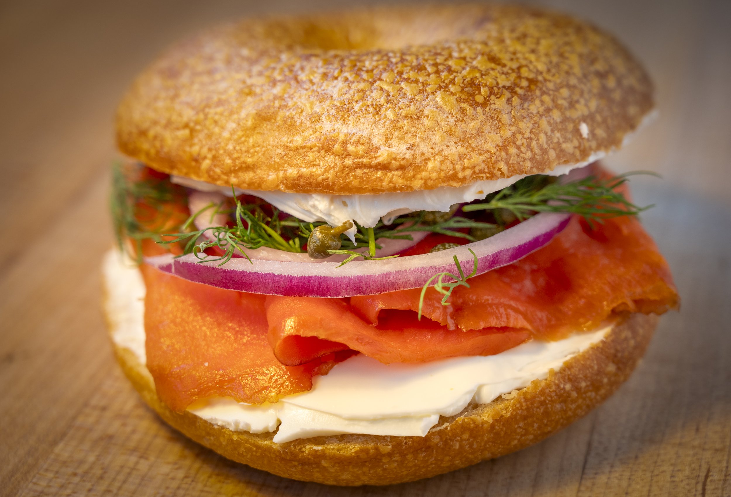 Close-up of a sandwich with a toasted bun, slices of smoked salmon, slices of red onion, fresh herbs, and a slice of white cheese.