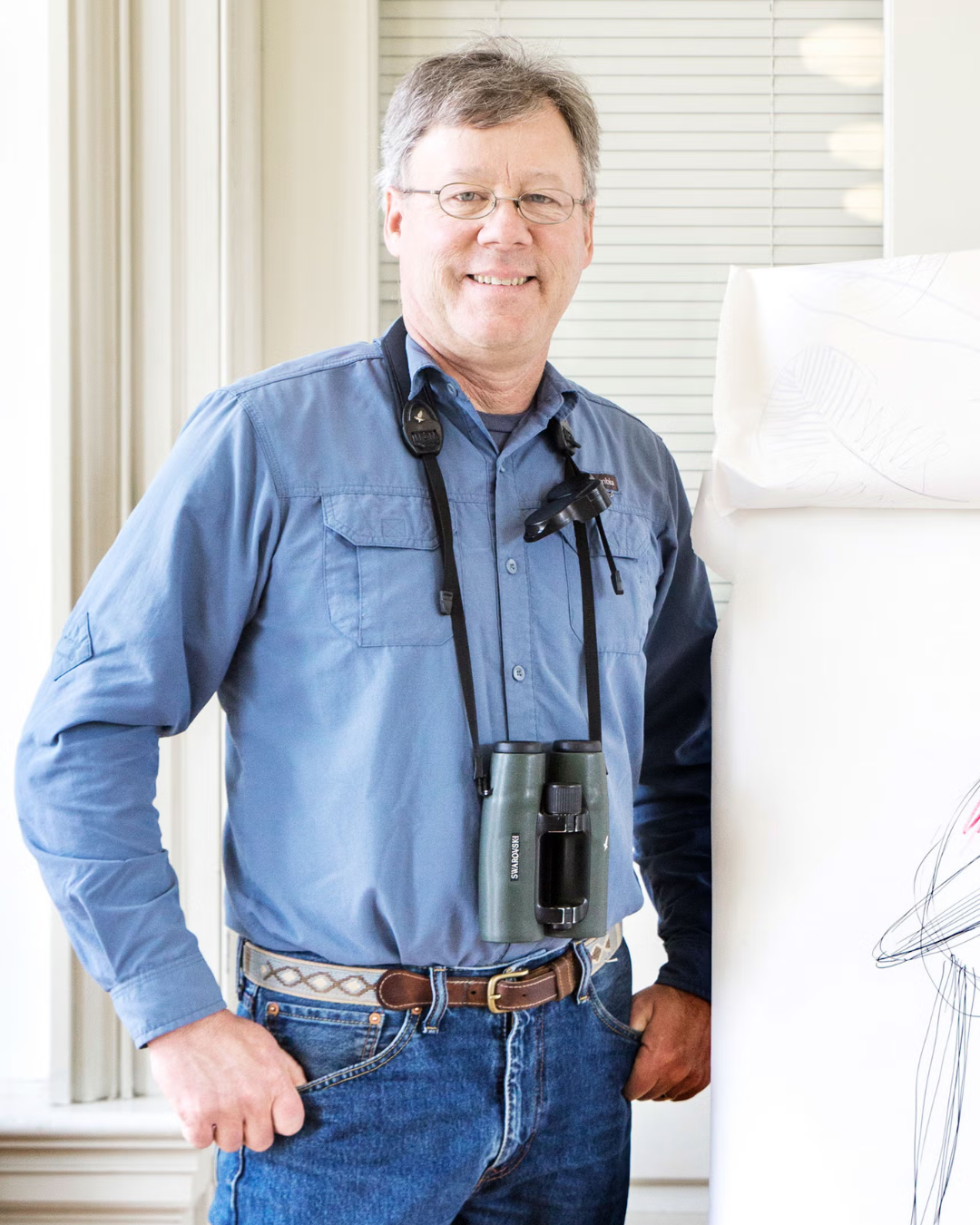 A man wearing glasses and a blue shirt stands indoors with binoculars hanging around his neck, smiling at the camera.